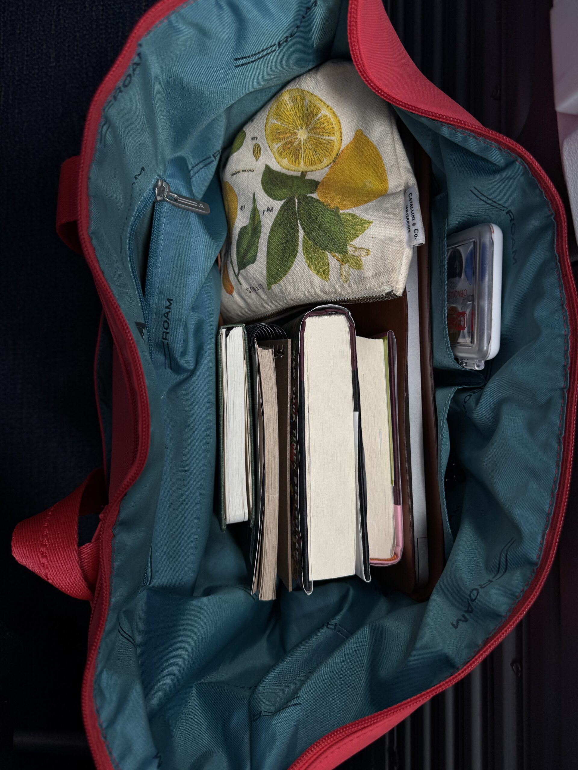 An open teal tote bag contains several books, a lemon-patterned pouch, a white tray, and a small clear case with items inside.