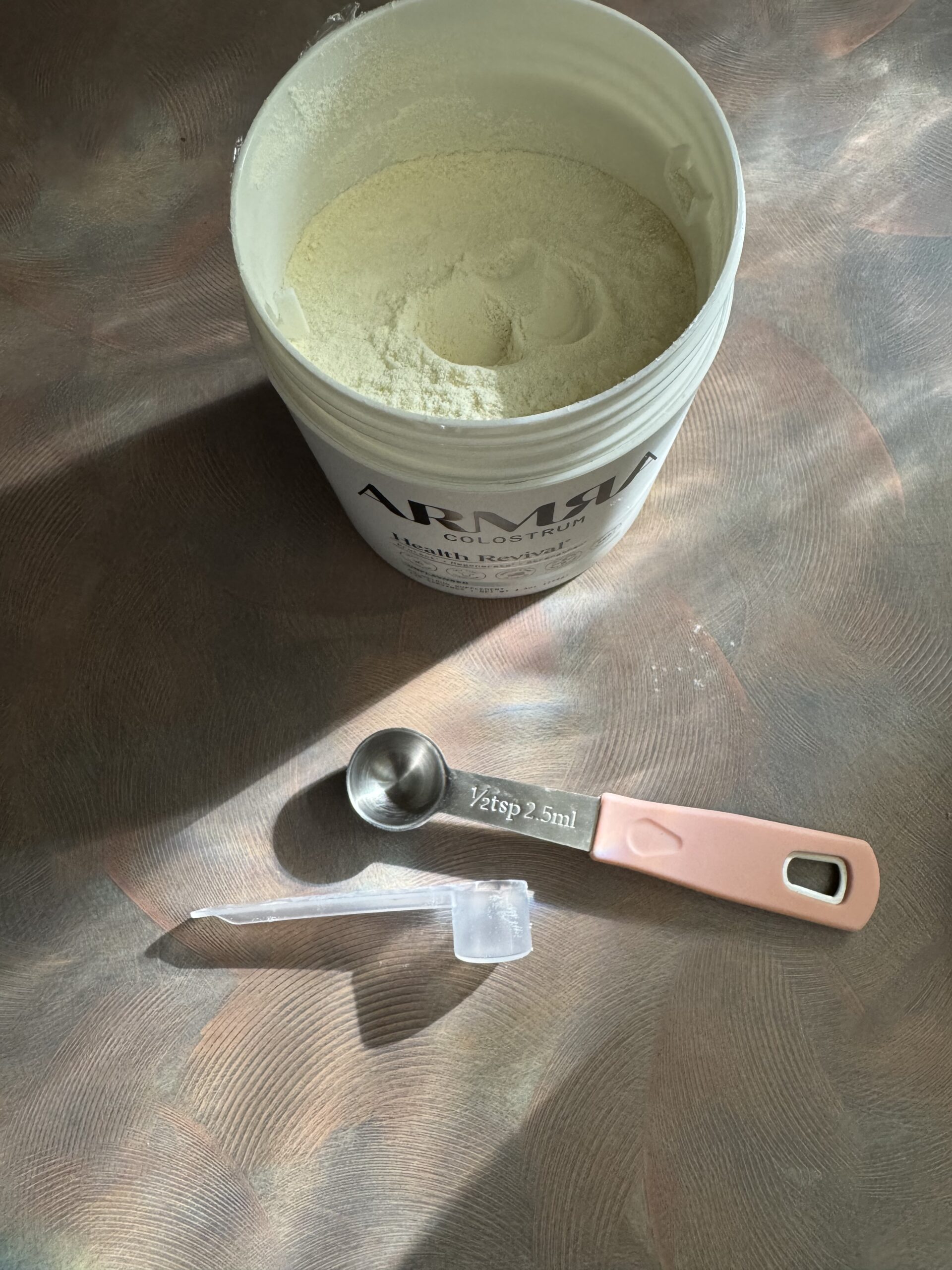 A container of powdered formula sits open next to a 2.5 ml measuring spoon and a clear plastic scoop on a reflective surface.