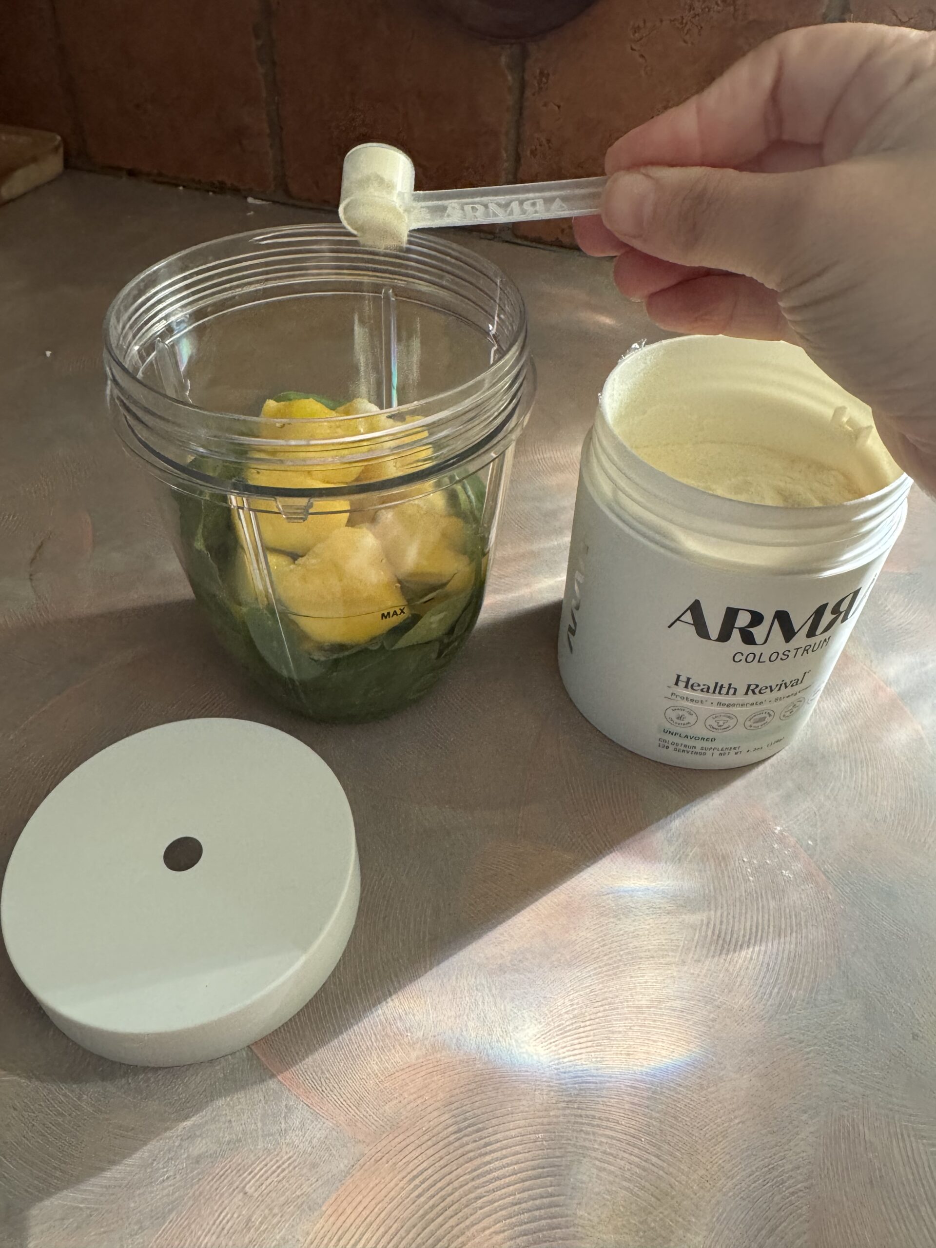 A hand adds a scoop of ARMRA colostrum powder to a blender cup containing greens and pineapple chunks, with the powder container and lid nearby on the counter.