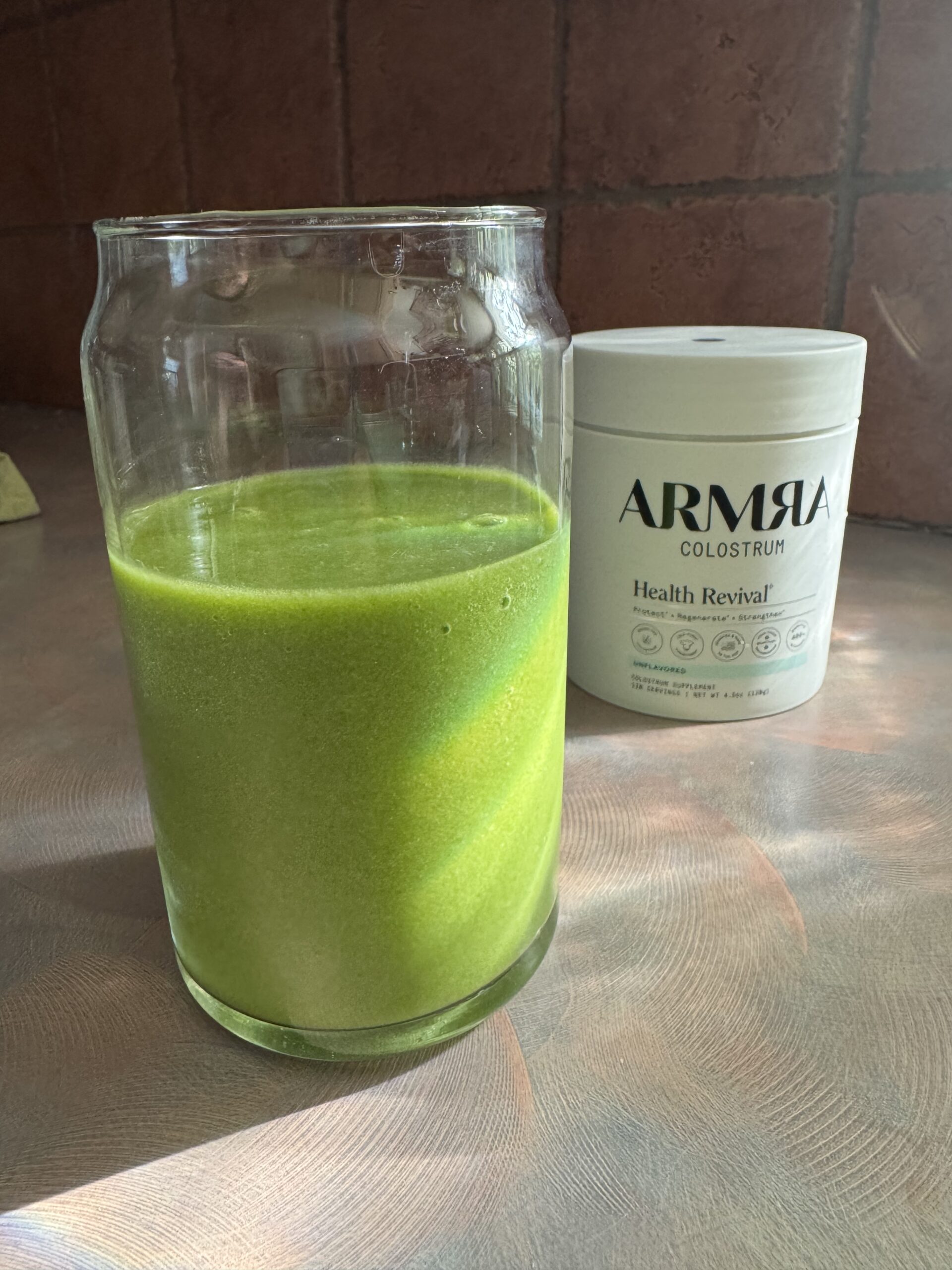 A glass of green smoothie sits on a counter, with a container labeled "ARMRA Colostrum Health Revival" in the background.