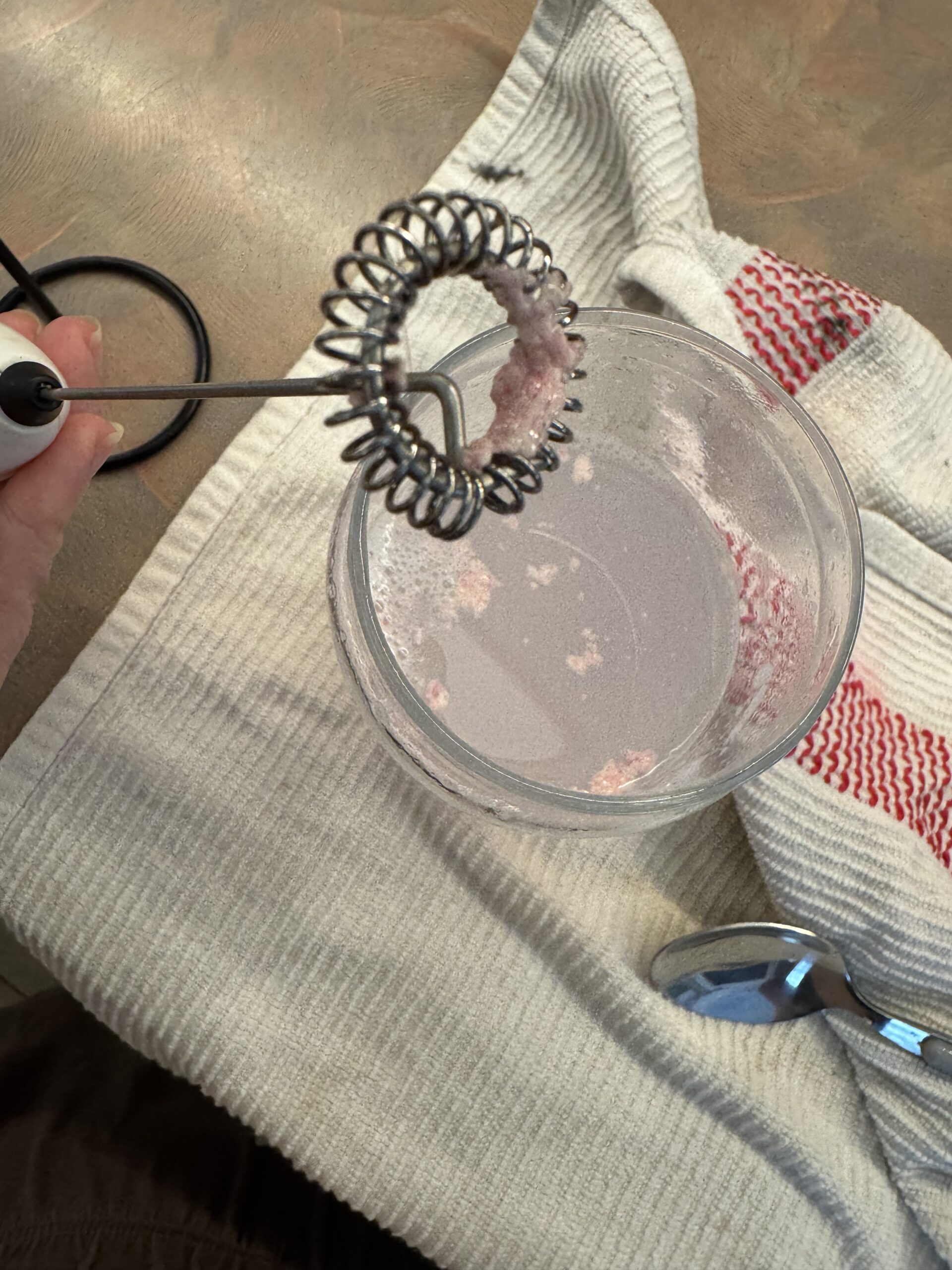 A hand holds a milk frother with pink residue above a glass of pink liquid, next to a spoon and white kitchen towels on a countertop.