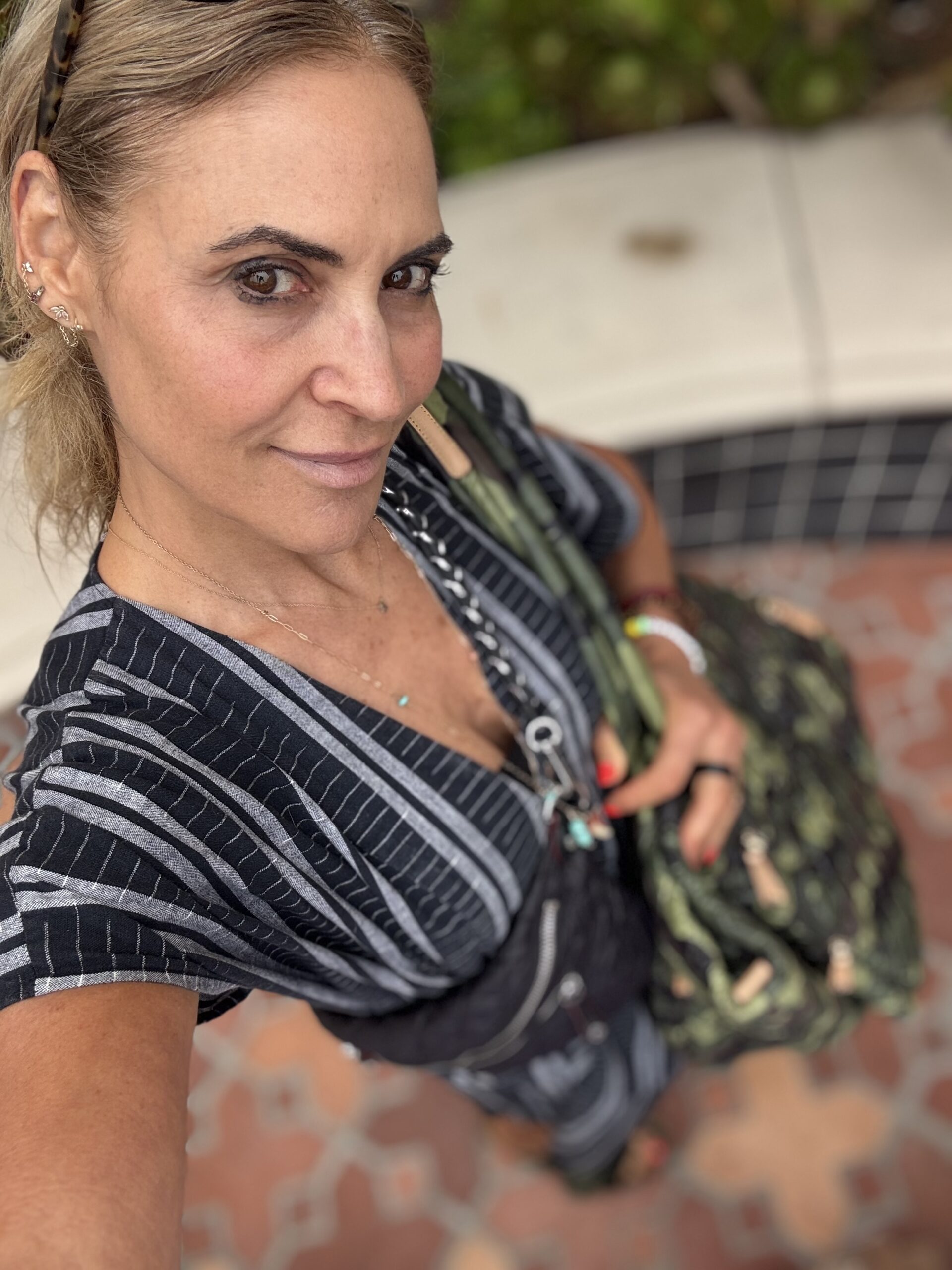 A person with light brown hair in a low ponytail wears a striped outfit and holds a green patterned bag, standing outdoors on a tiled surface.