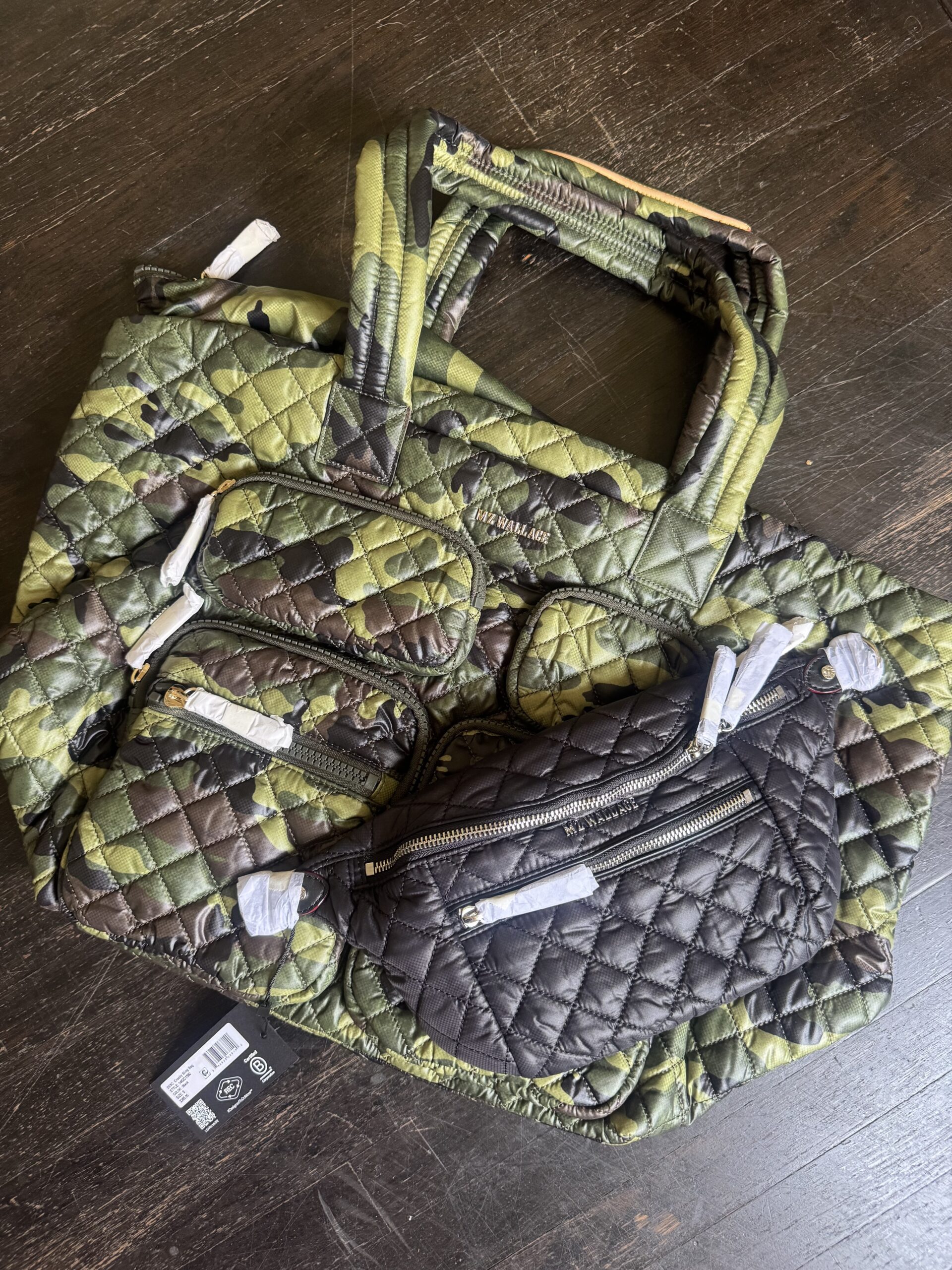 Two quilted bags, one green camouflage tote and one black crossbody, are placed on a dark wooden floor. Both bags have multiple zippered compartments and tags attached.