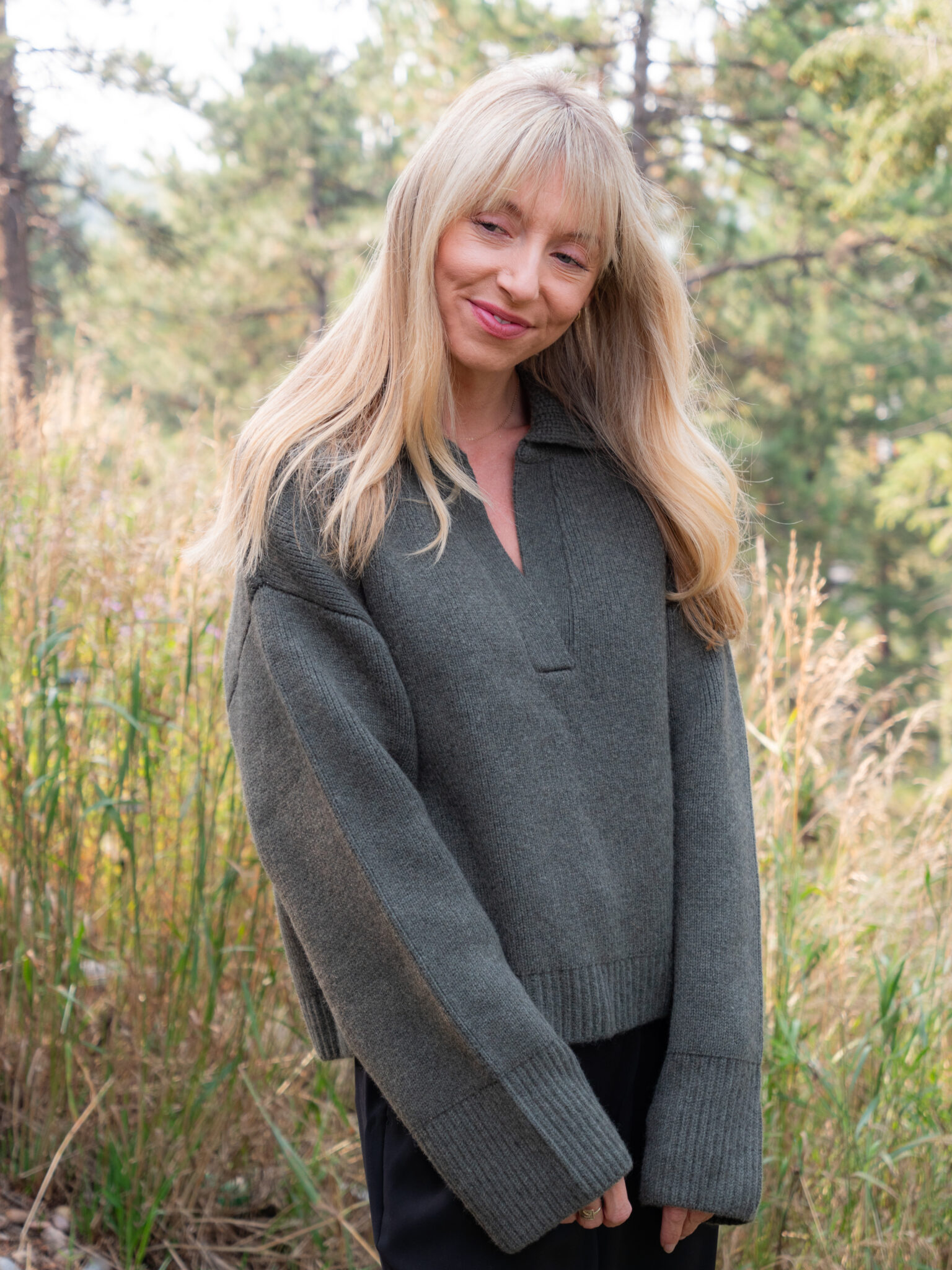 A woman with long blonde hair wearing a dark green sweater stands outdoors in front of tall grass and trees.