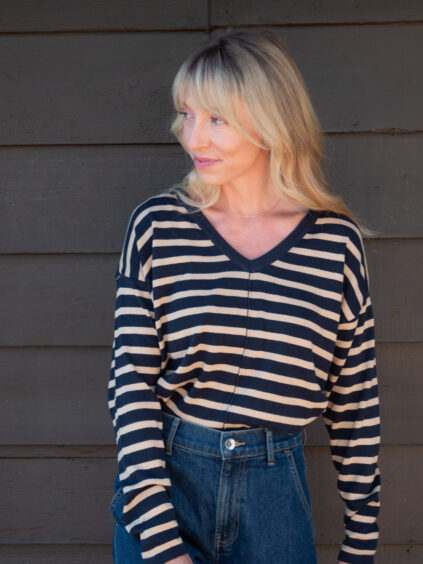 A woman with blonde hair wearing a black and beige striped long-sleeve shirt and high-waisted blue jeans stands in front of a dark wooden wall, looking to the side.