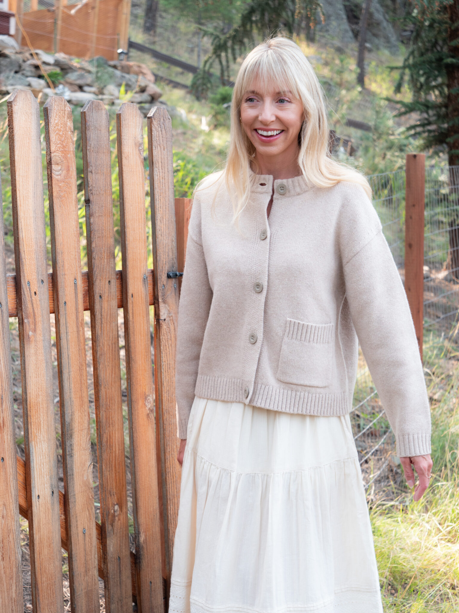 A woman with long blonde hair stands outdoors by a wooden fence, wearing a beige button-up sweater and a white skirt. Trees and greenery are visible in the background.