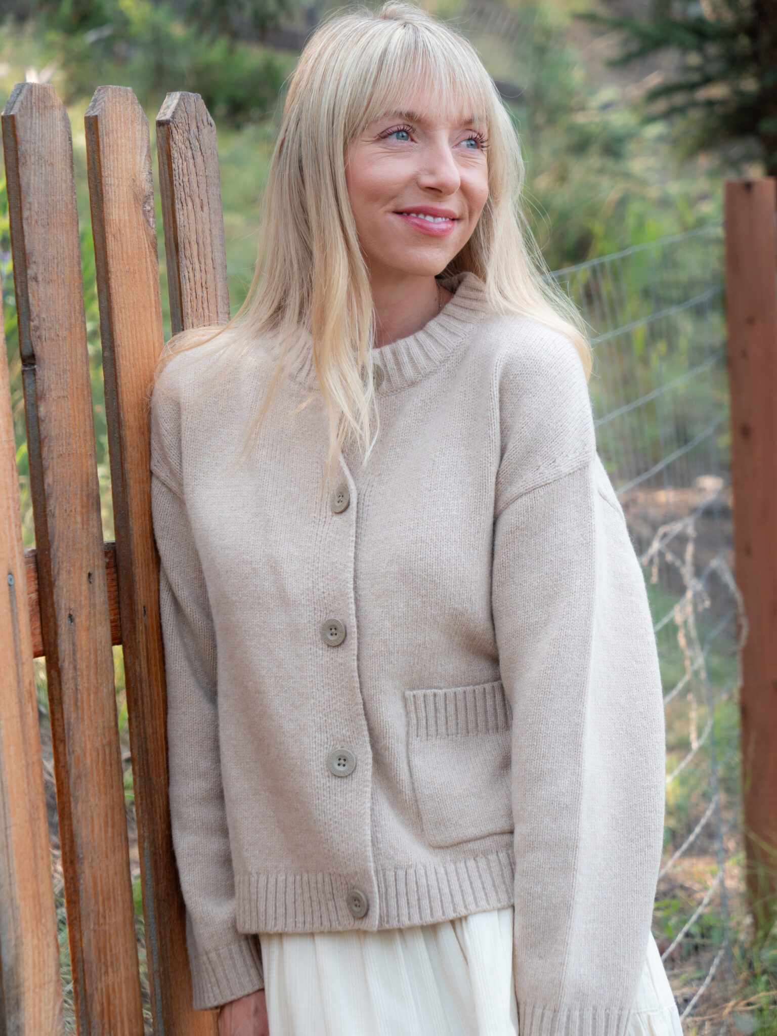 A woman with long blonde hair, wearing a beige buttoned sweater, stands outdoors near a wooden fence, smiling slightly.