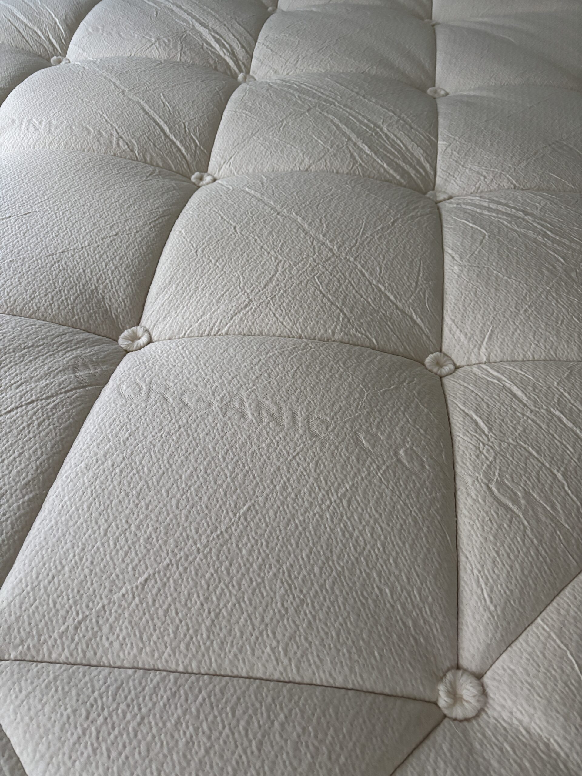 Close-up of a textured, light-colored, tufted mattress surface with visible stitching and button-like indentations.