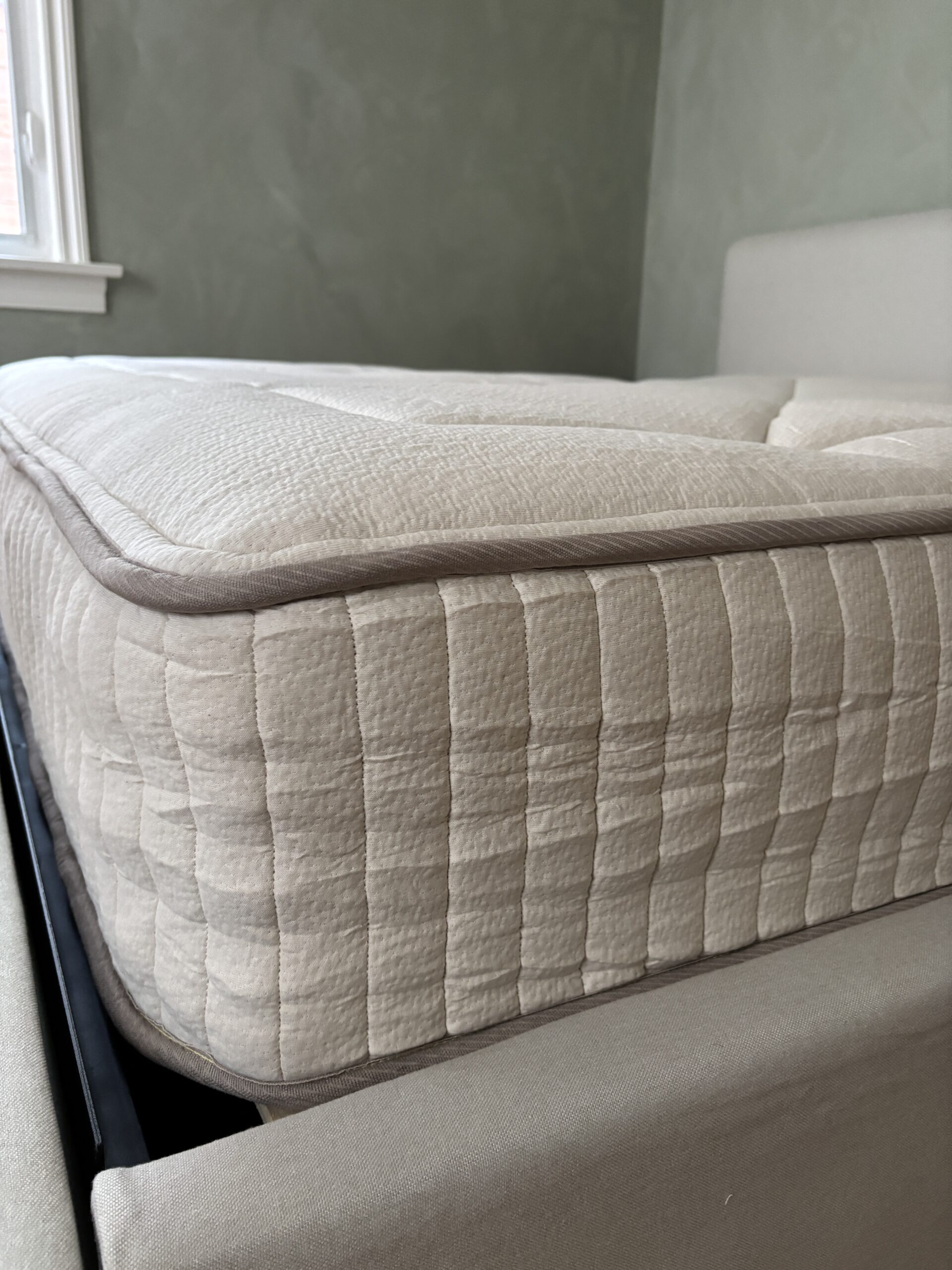 A close-up view of a beige, quilted mattress on a bed frame in a room with light-colored walls and a window in the background.