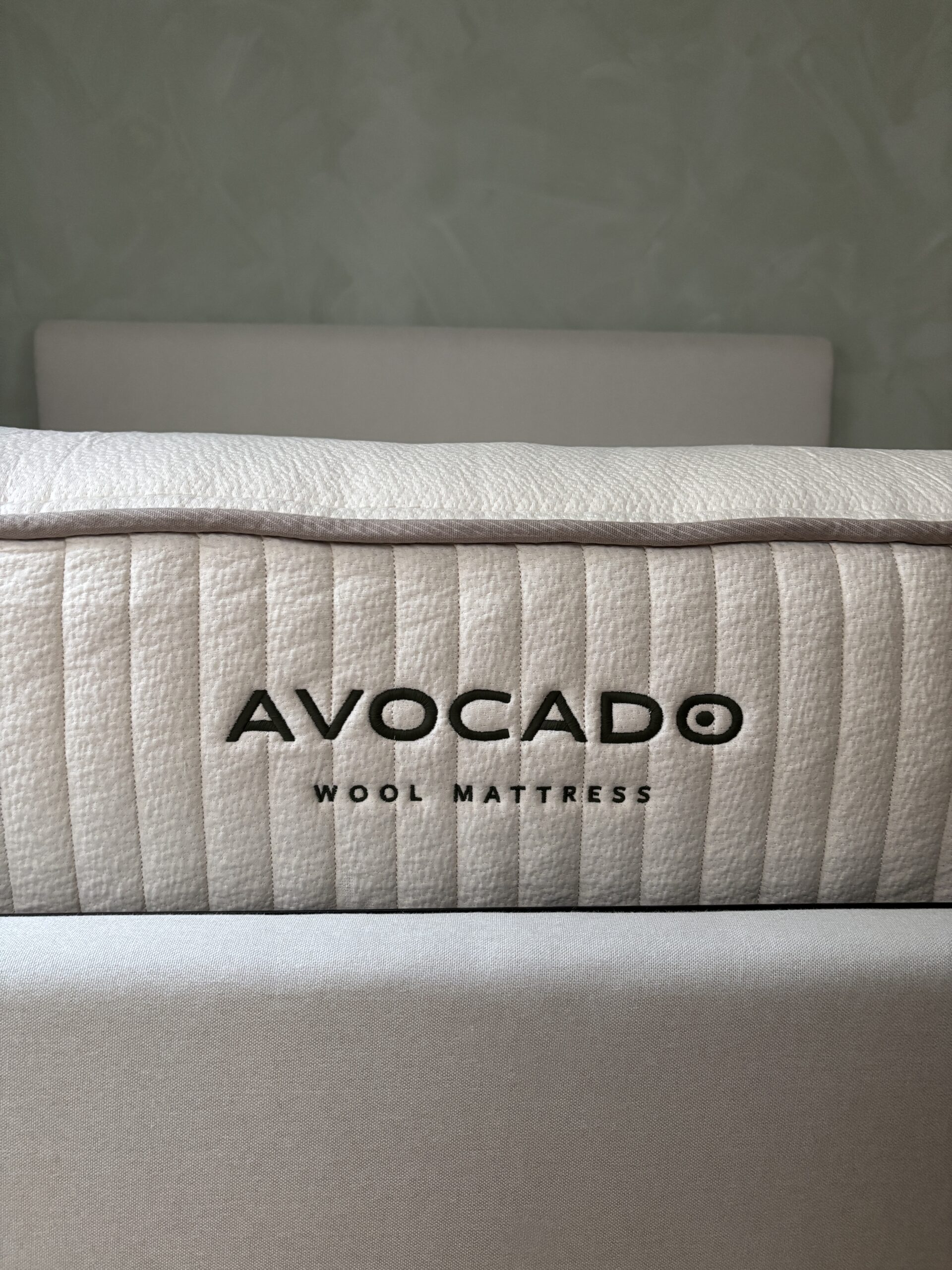 Close-up of a mattress with the brand name "Avocado" and the words "Wool Mattress" embroidered on the side, positioned against a light-colored headboard.