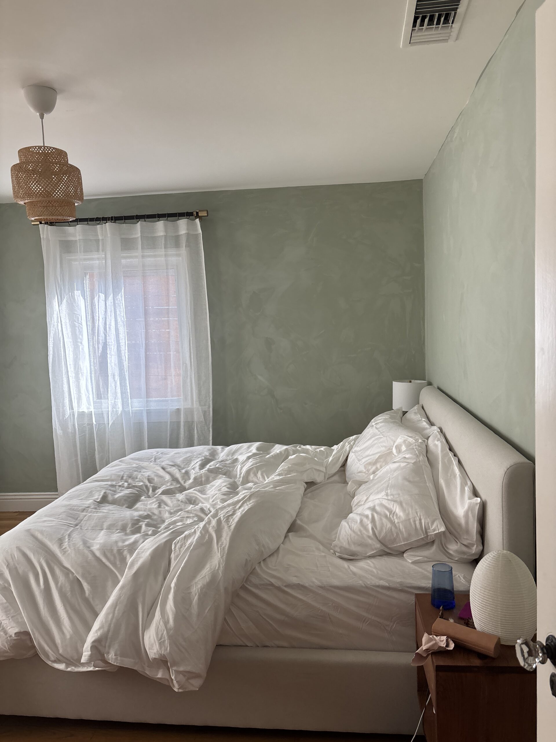 A disheveled bed with white bedding sits next to a wooden nightstand in a softly lit bedroom with pale green walls and a window with sheer curtains.