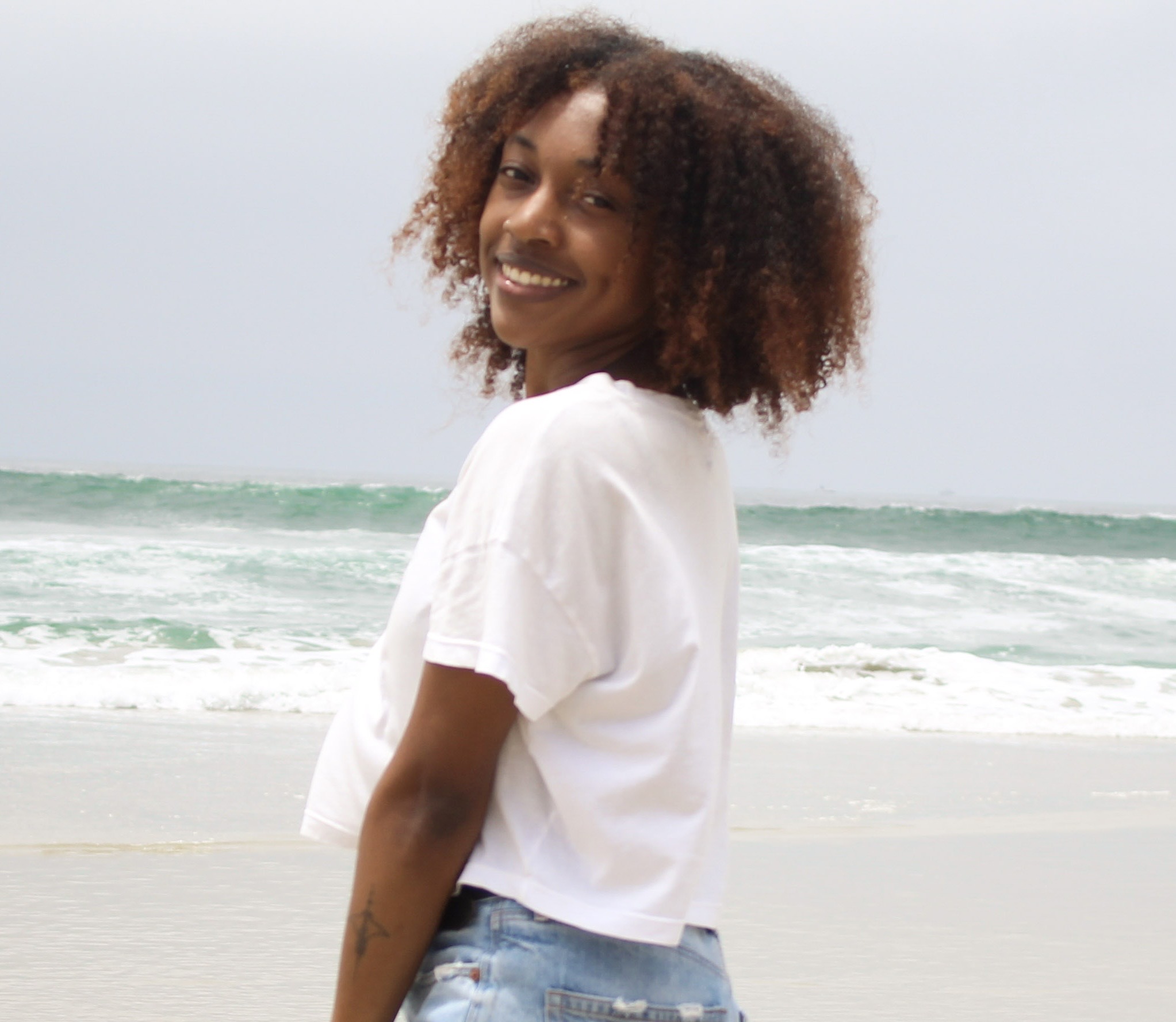 A person with curly hair, wearing a white t-shirt and denim shorts, stands on a beach with waves in the background, smiling at the camera.