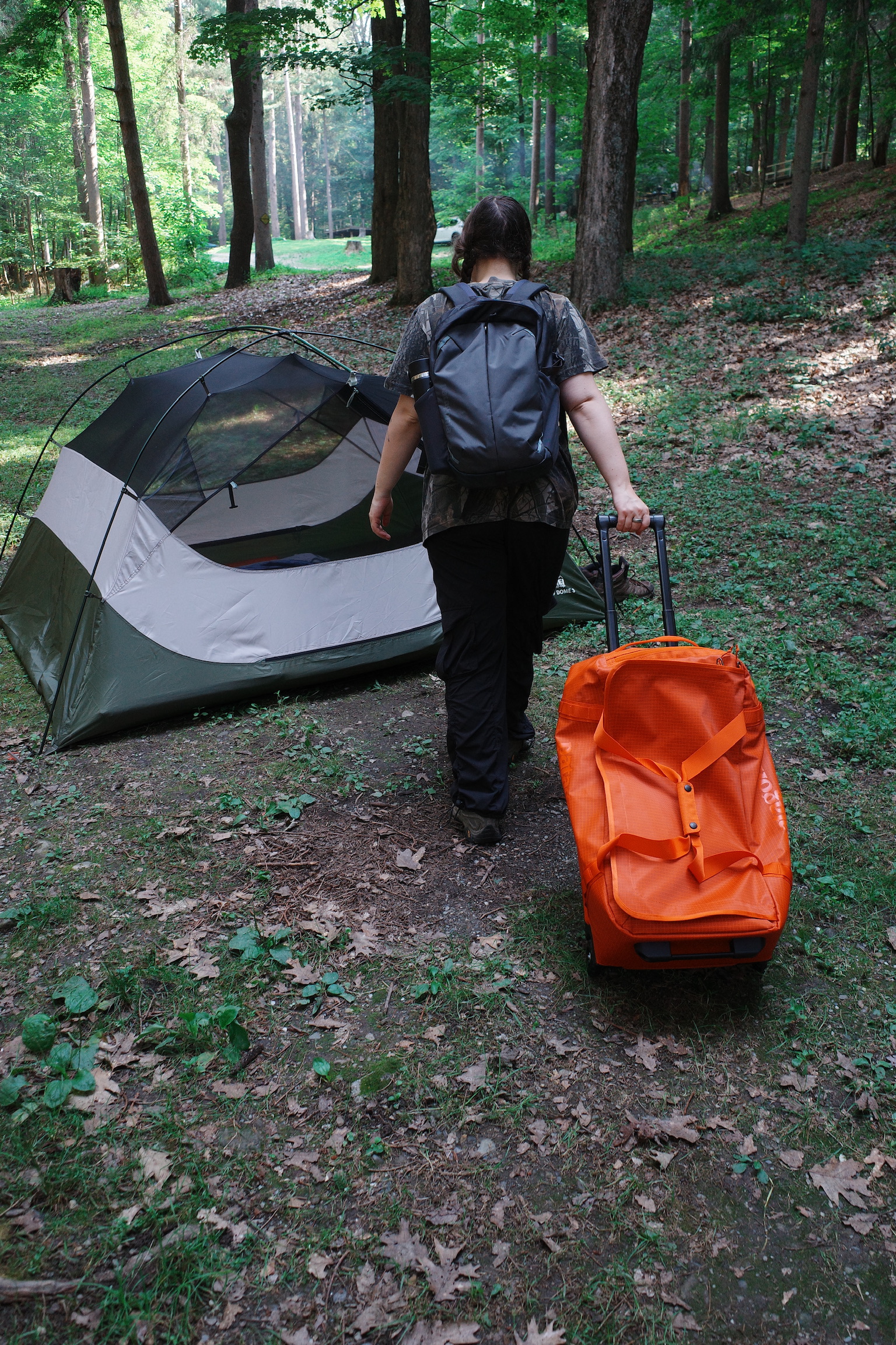 A person with a backpack pulls an orange suitcase toward a tent set up in a wooded campsite.