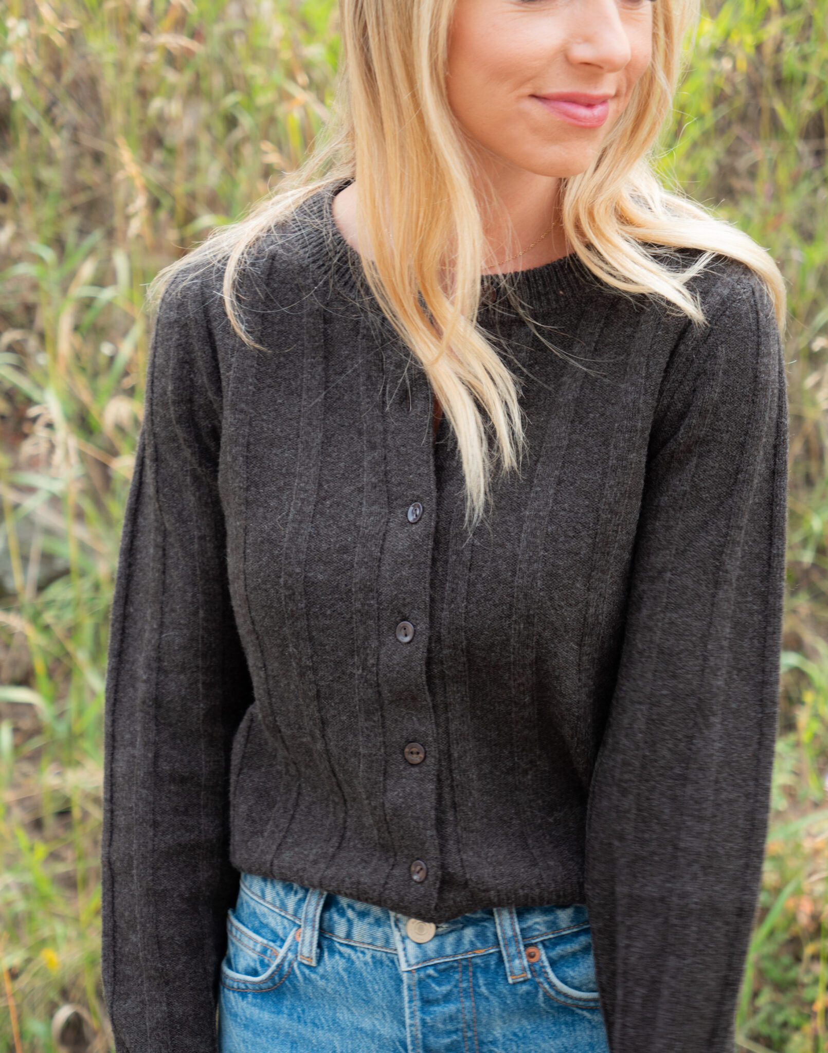 Woman in a dark ribbed button-up sweater and blue jeans standing outdoors with tall grass in the background.