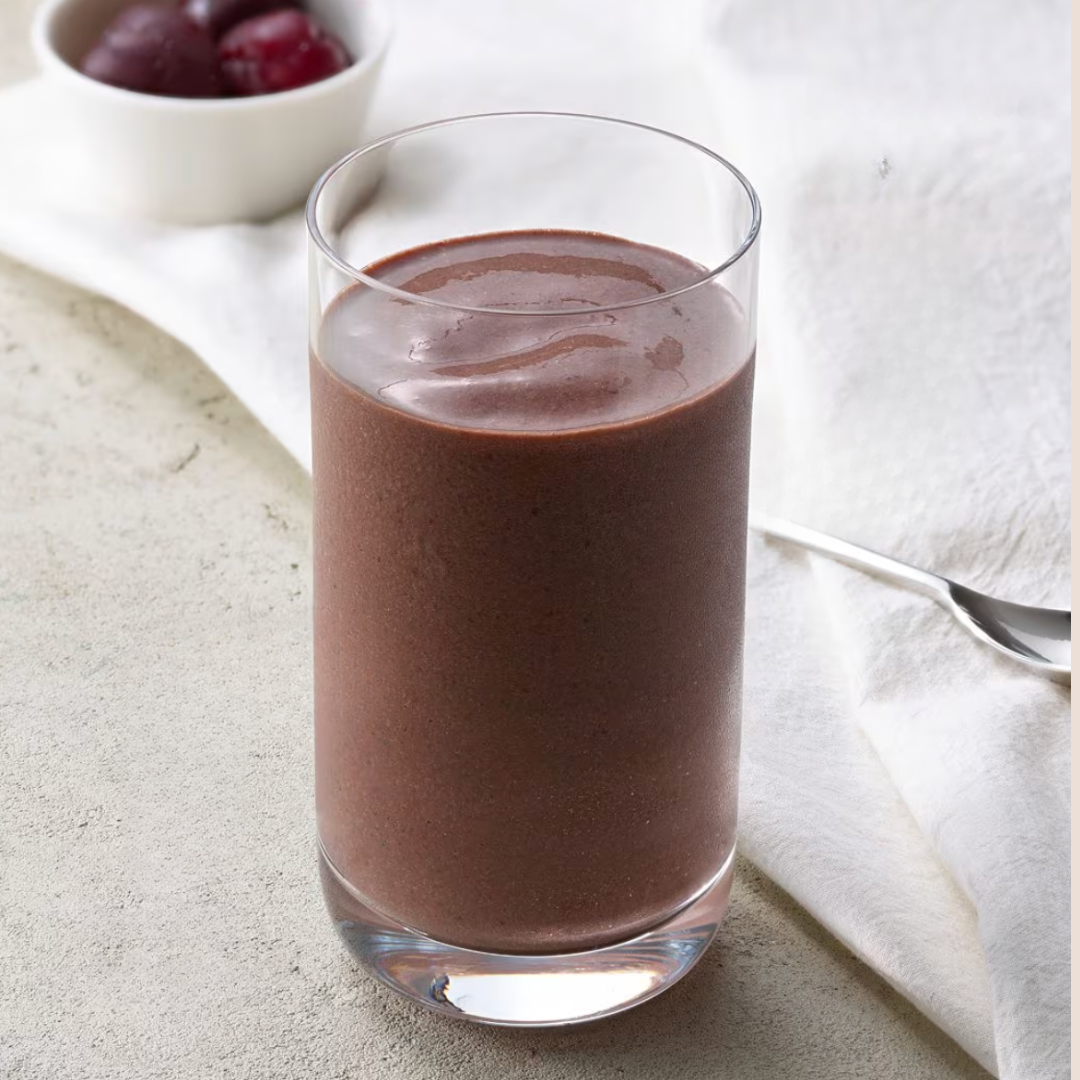 A glass filled with a thick chocolate smoothie sits on a light surface; a spoon and a small bowl of cherries are in the background.