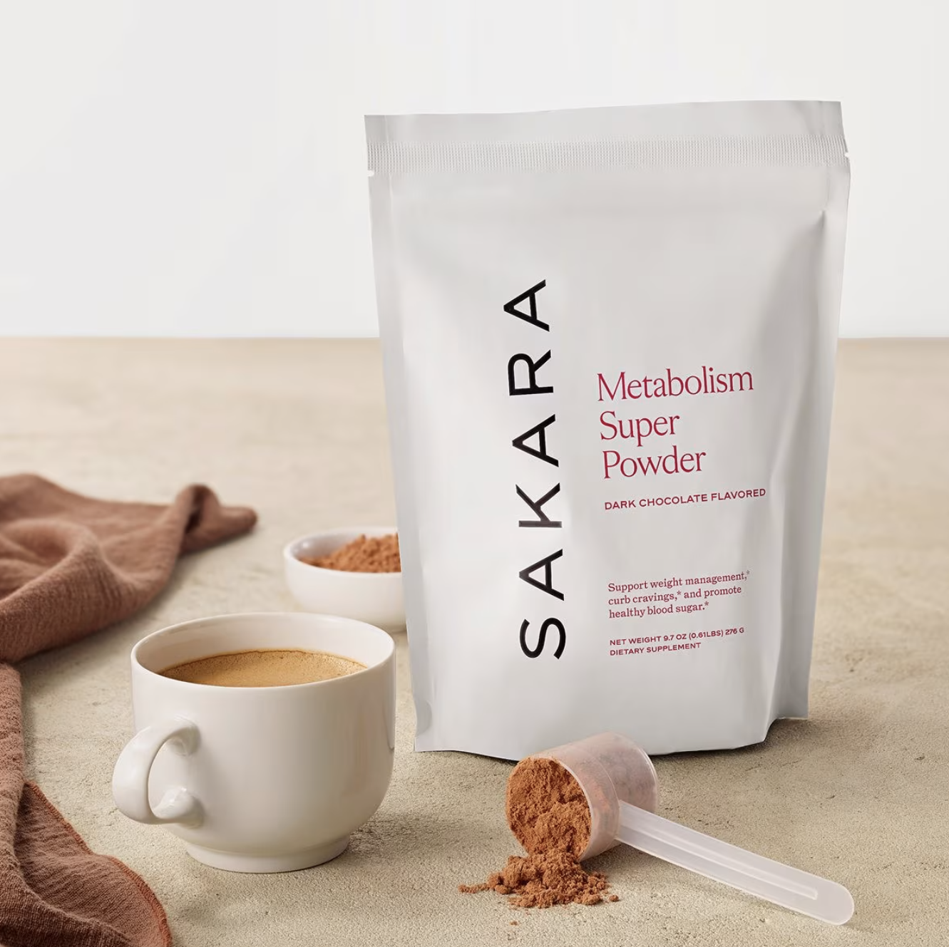 A bag of Sakara Metabolism Super Powder stands upright next to a white mug of prepared drink, a scoop of powder, and a brown cloth on a beige surface.