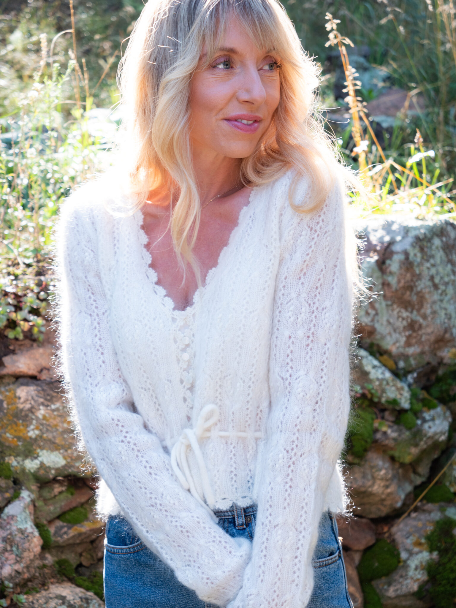A woman with long blonde hair wearing a white knitted sweater and blue jeans stands outdoors in sunlight, with a stone wall and greenery in the background.