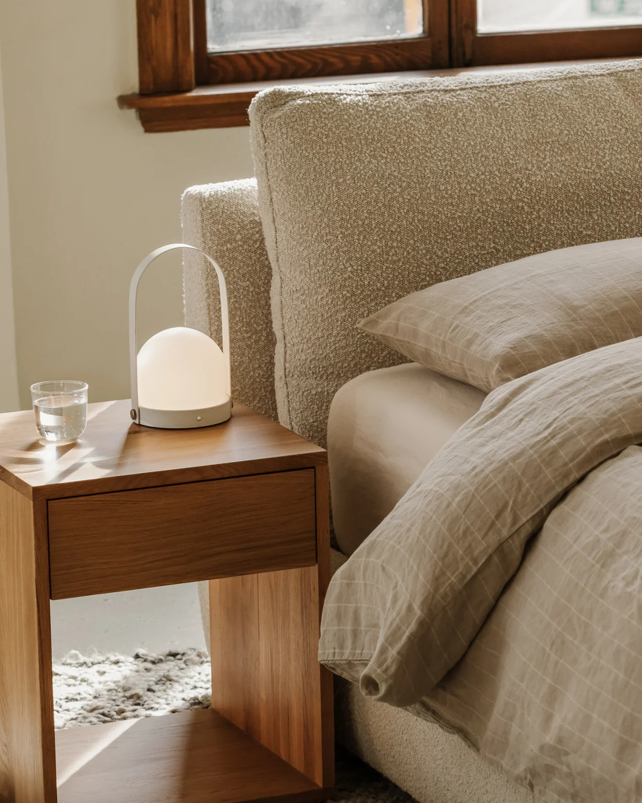 A wooden bedside table with a glass of water and a modern lamp beside a bed with beige linens in a softly lit room.
