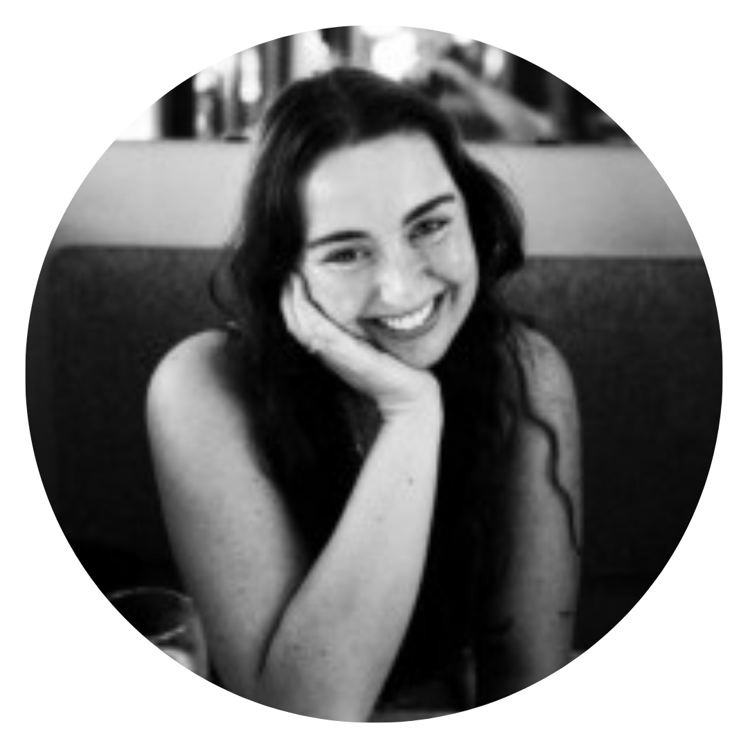 A woman with long dark hair smiles and rests her chin on her hand while seated at a table. The photo is black and white and framed in a circular crop.
