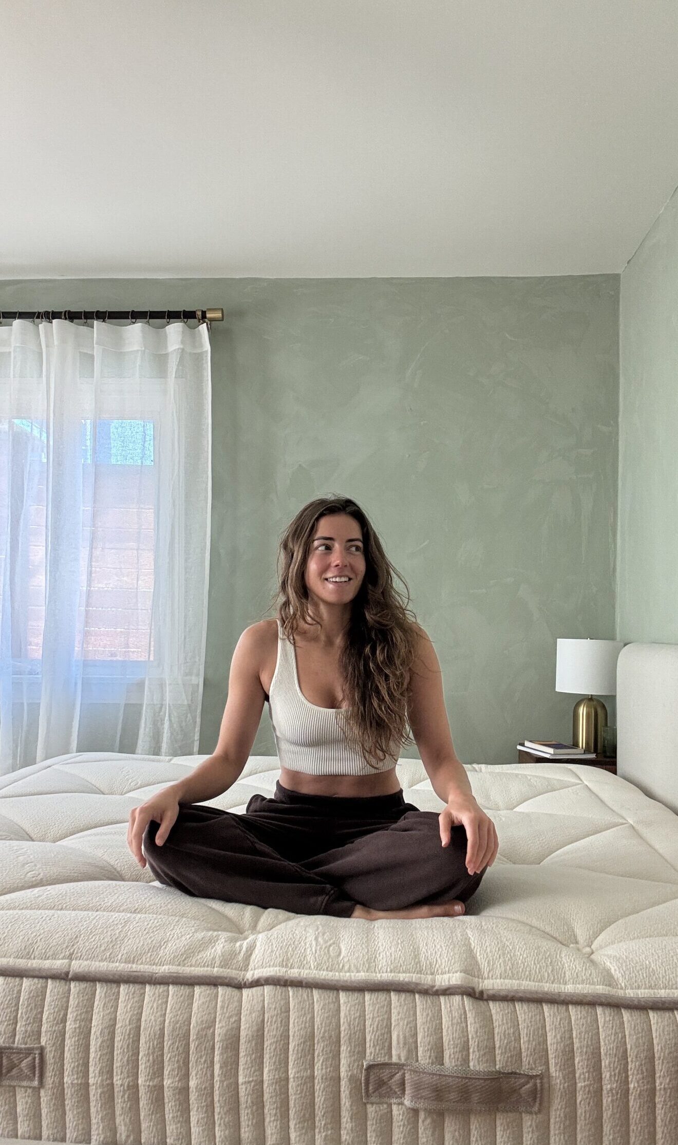 A woman sits cross-legged on a large, unmade bed in a bright bedroom with green walls and sheer curtains.