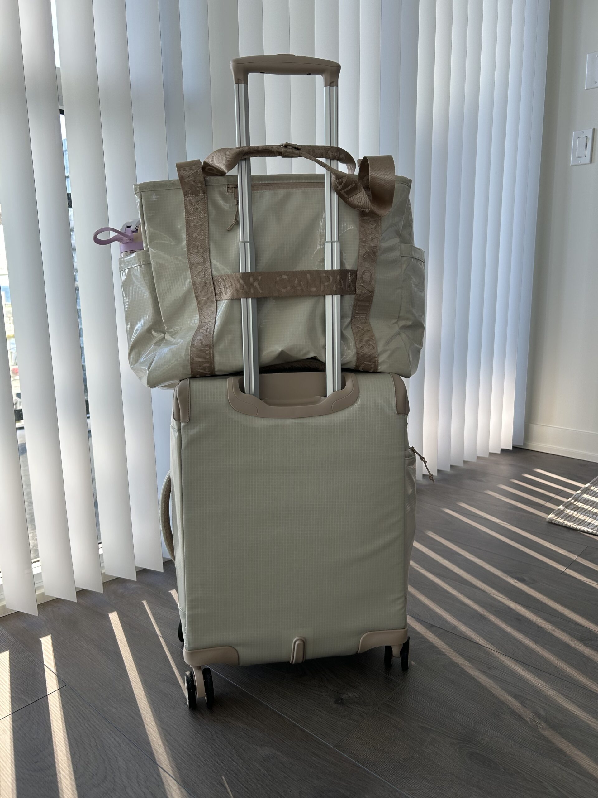 A beige rolling suitcase with a matching tote bag stacked on top, positioned in front of vertical blinds with sunlight casting shadows on the floor.