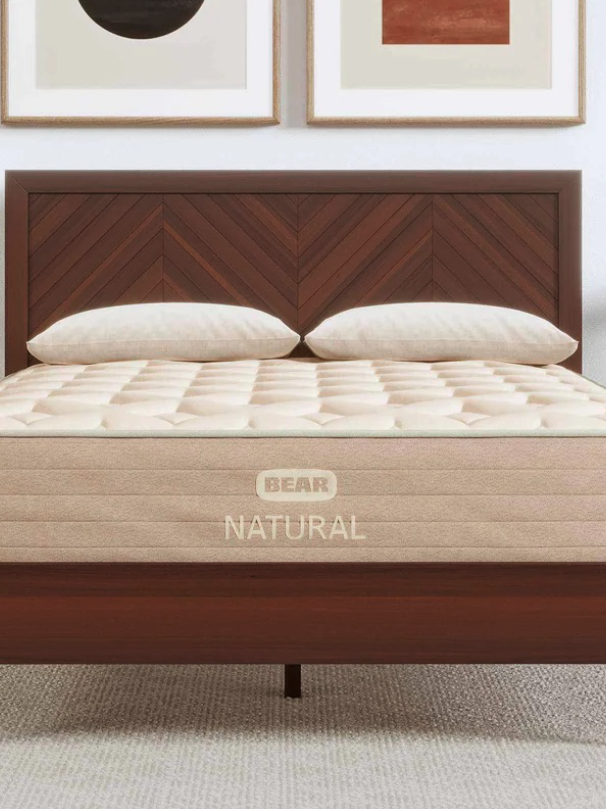 A neatly made bed with a wooden headboard, two pillows, and a mattress labeled "Bear Natural" in a minimalist bedroom with framed art on the wall.