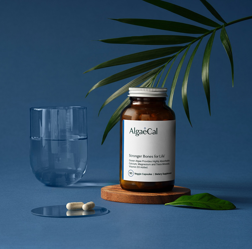 A bottle of AlgaeCal supplements, two capsules, a glass of water, a small mirror, and a green leaf are arranged on a blue surface with a palm frond in the background.