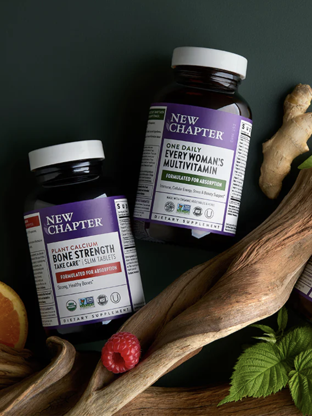 Three New Chapter supplement bottles are arranged with ginger root, green leaves, an orange slice, and a raspberry on a dark surface.