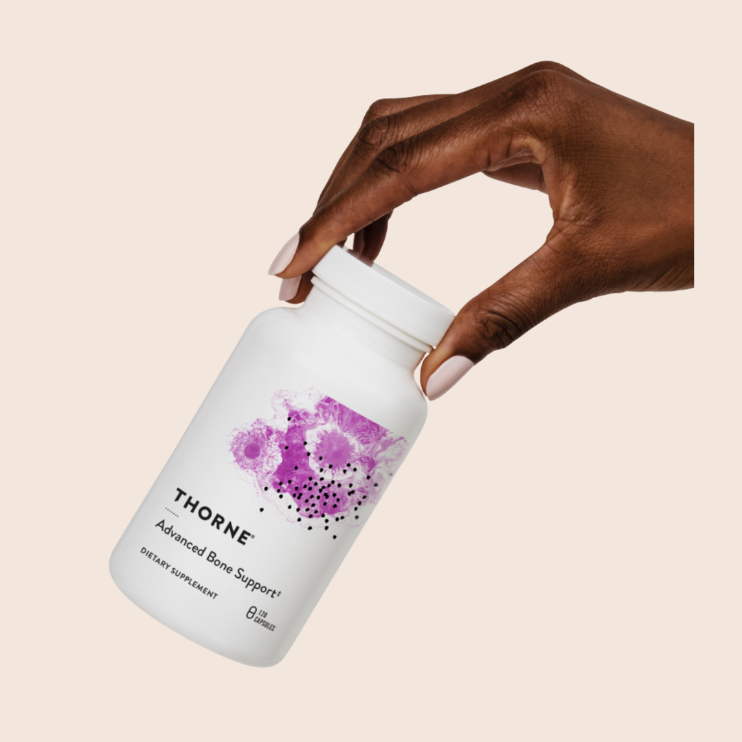A hand holds a white bottle labeled "Thorne Advanced Bone Support" with a purple abstract design on the front.