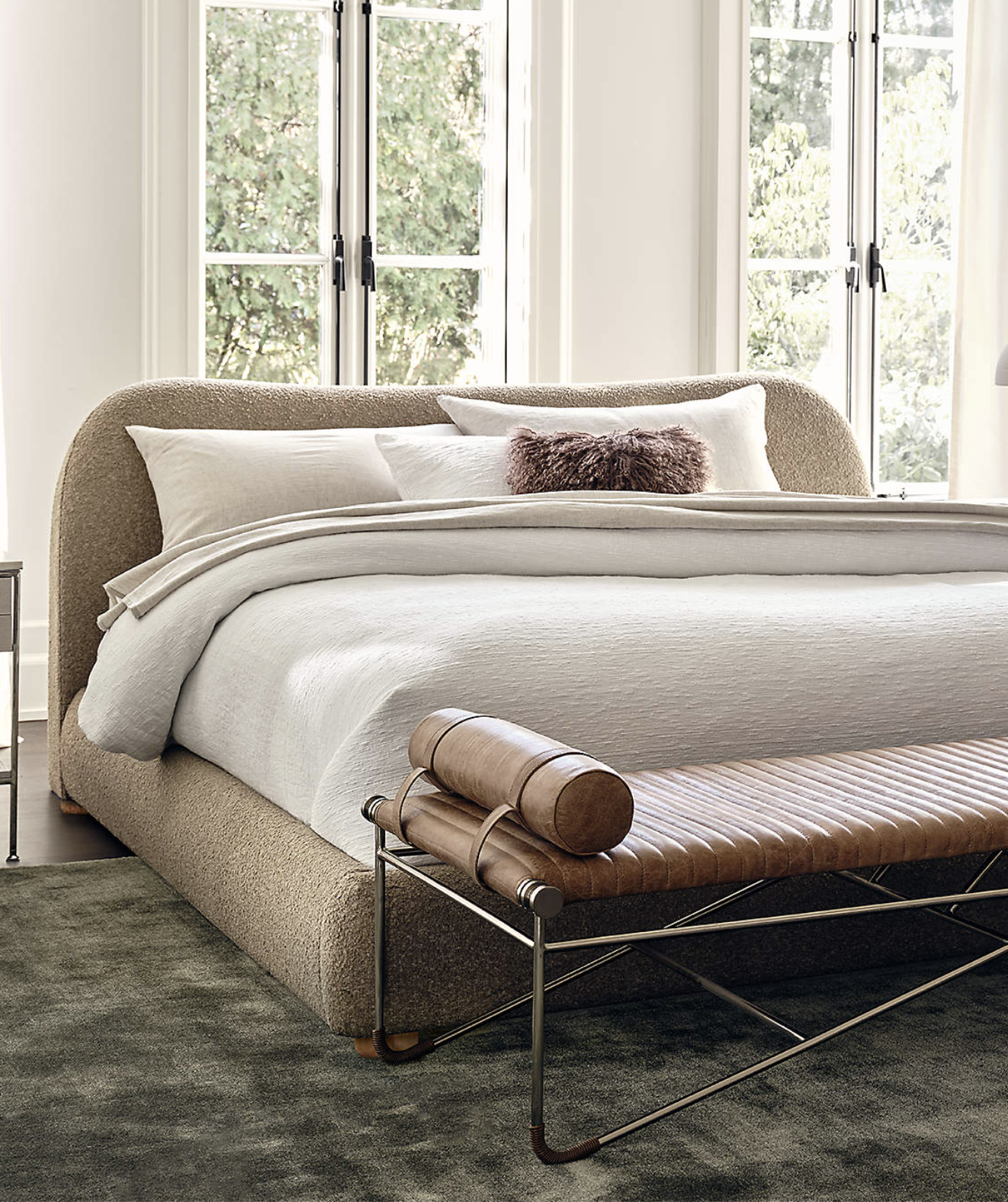 A neatly made bed with white bedding and a textured beige headboard, accompanied by a brown leather bench at the foot, sits in front of tall windows with greenery outside.