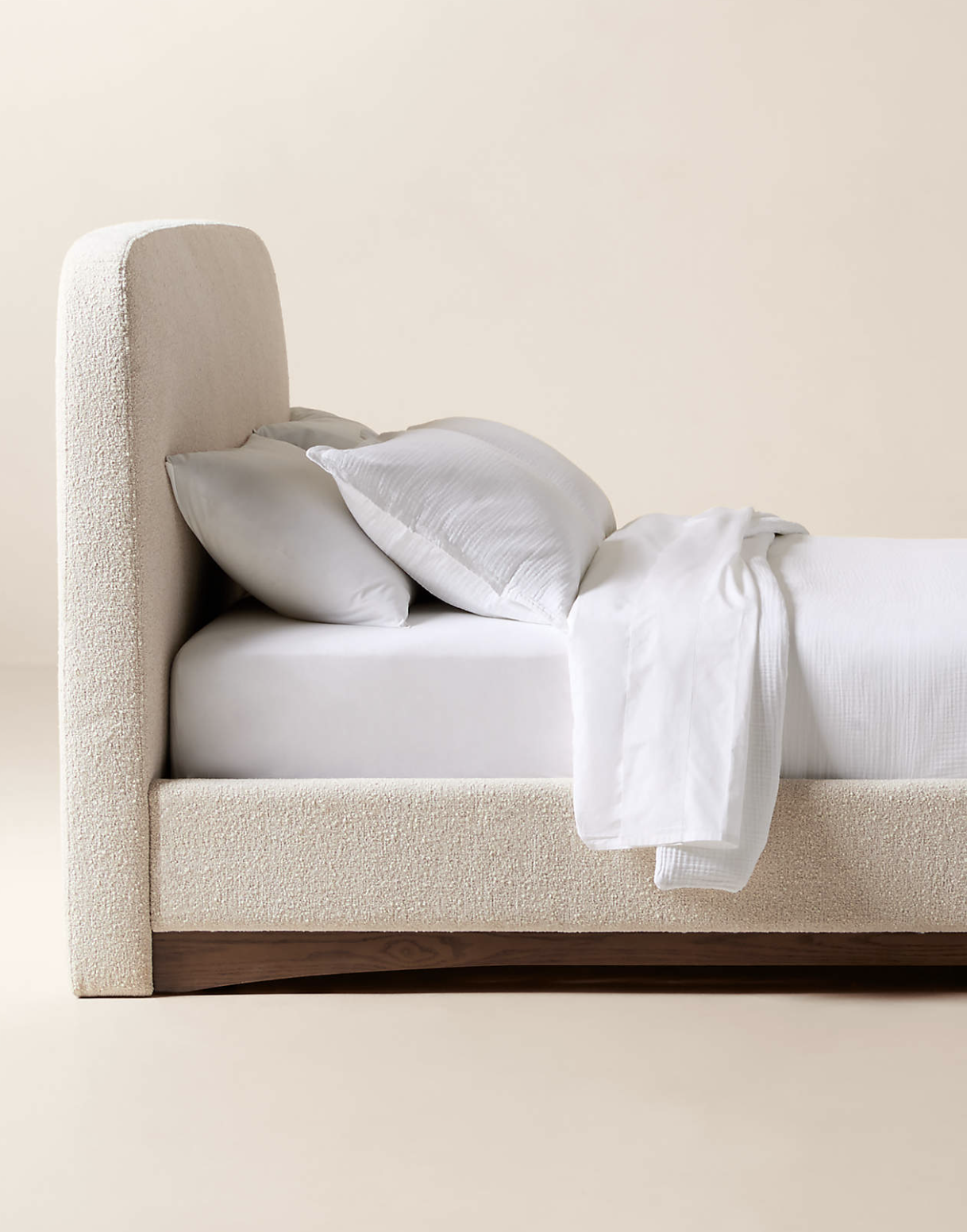 A side view of a modern upholstered bed with white bedding, pillows, and a wooden base against a plain off-white background.