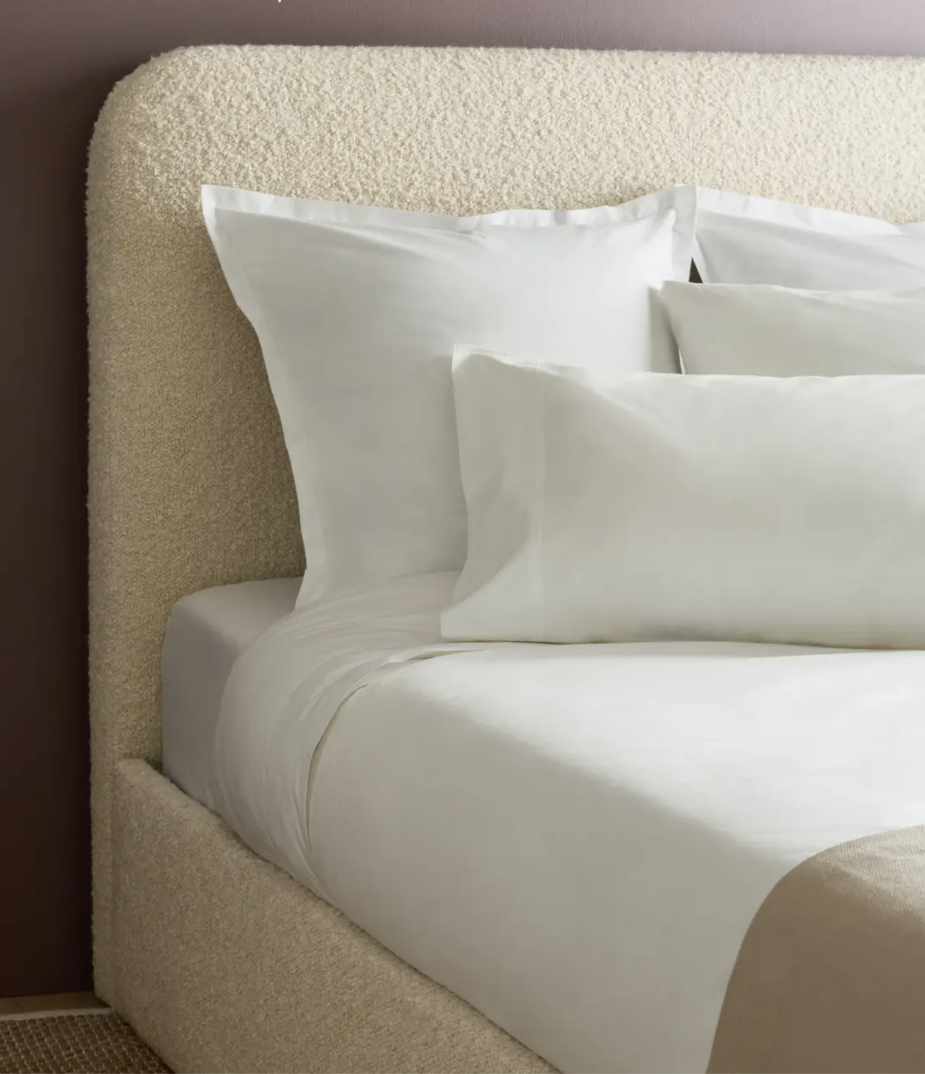 A neatly made bed with white sheets, pillowcases, and shams, featuring a textured upholstered headboard and a beige blanket at the foot of the bed.