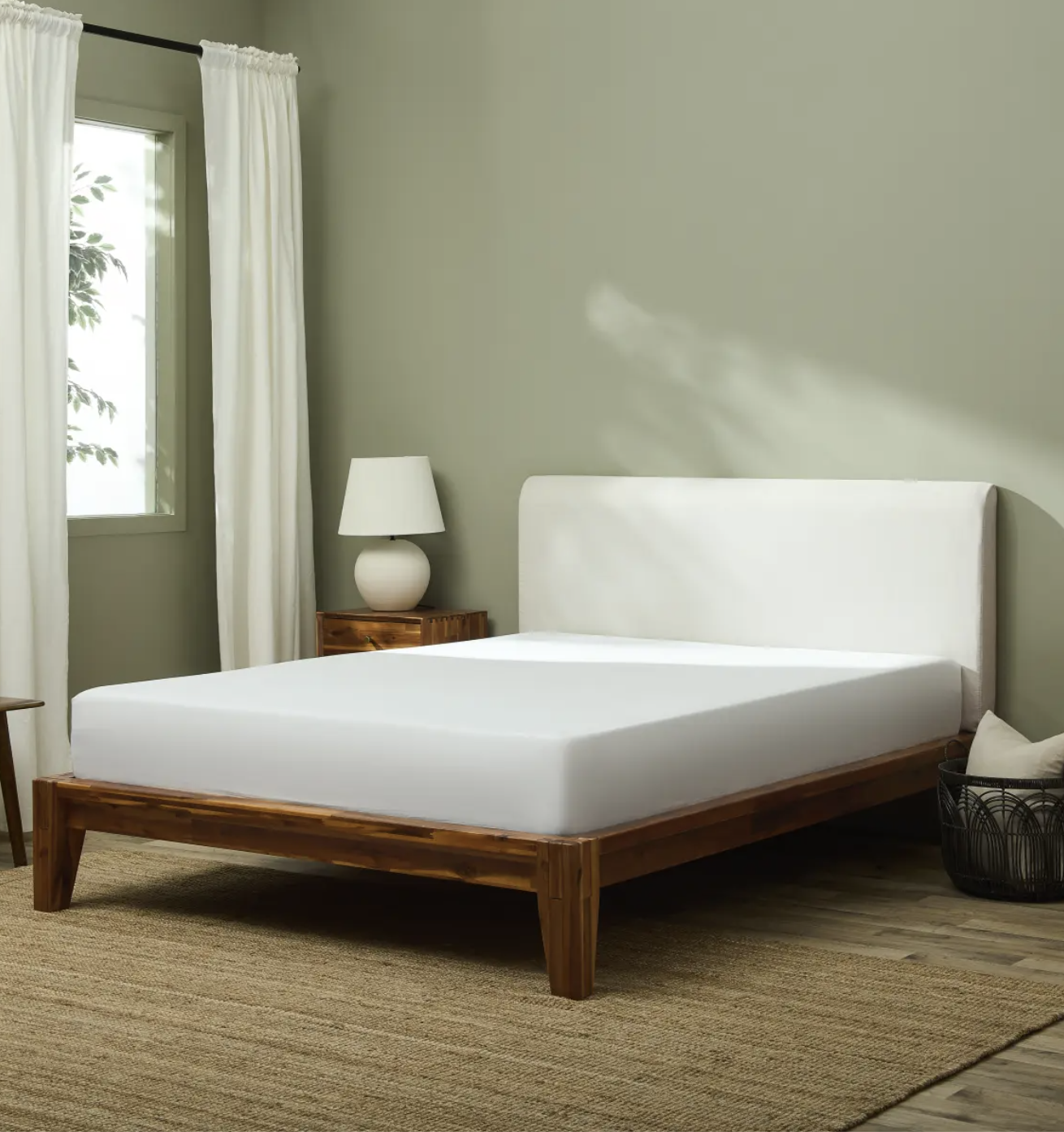 A neatly made bed with a wooden frame and white bedding stands in a simple bedroom with a bedside table, lamp, basket, and window with white curtains.