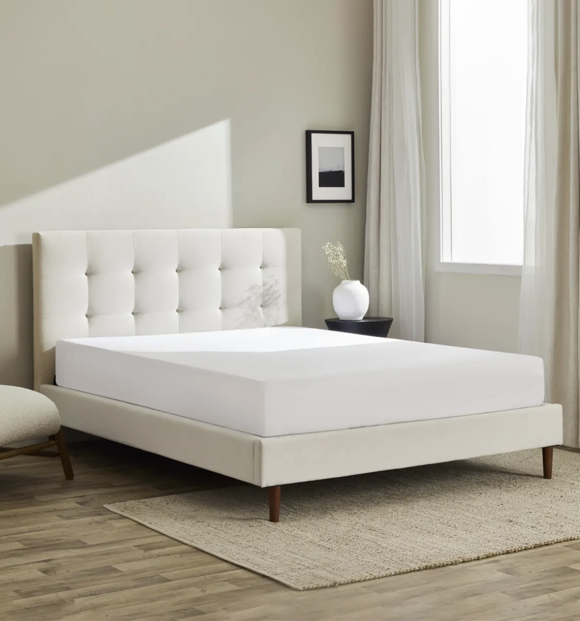 A modern bedroom with a white upholstered bed, white mattress, beige rug, light wood floor, chair, framed picture, vase on a table, and a large window with white curtains.