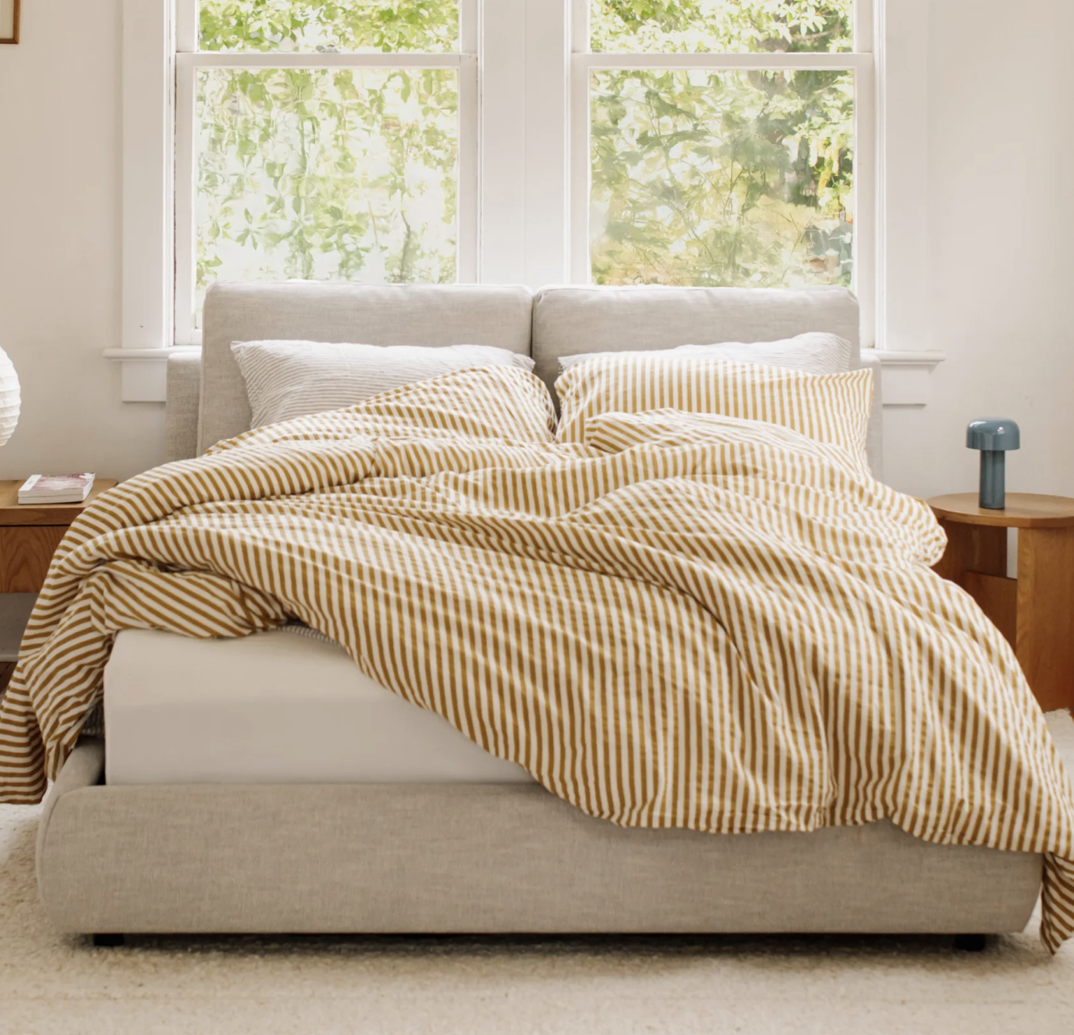 A neatly made bed with yellow and white striped bedding is positioned in front of two large windows with greenery visible outside. A nightstand and lamp are on either side of the bed.