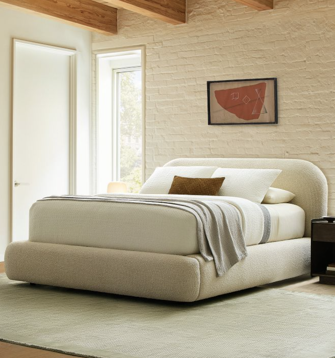 A modern bedroom with a light upholstered bed, gray throw blanket, brown accent pillow, nightstand, framed abstract art, and large windows on a light area rug.