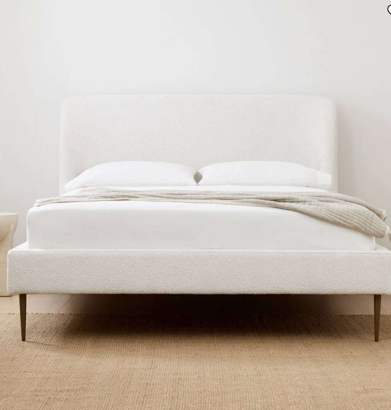 A minimalist bed with a white upholstered headboard, white bedding, and a light blanket, set on a woven rug against a plain white wall.
