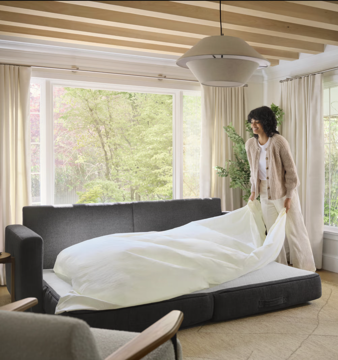 A person stands by a dark gray sofa bed, arranging a white duvet in a bright living room with large windows and light curtains.