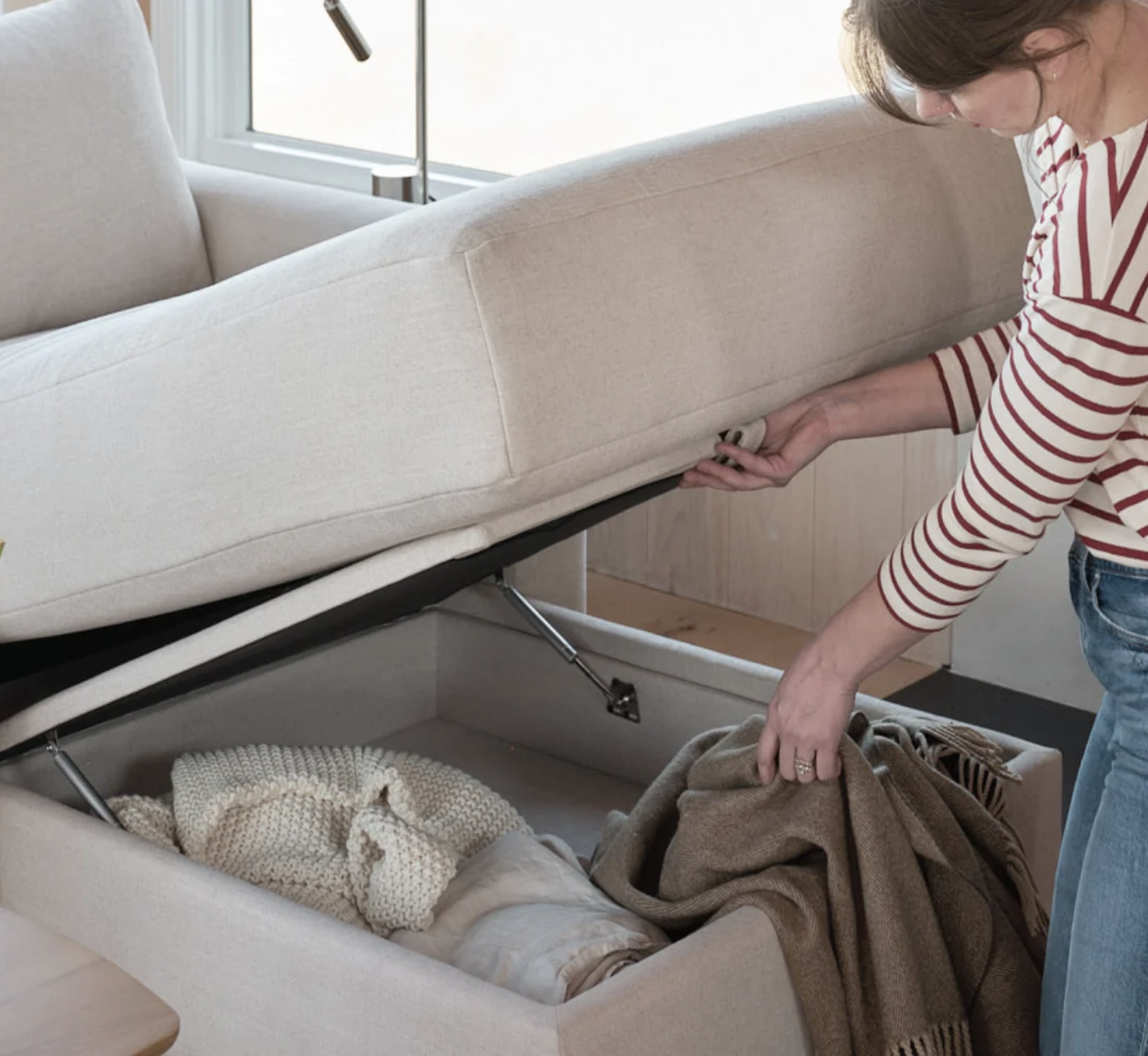 A person lifts the seat of a beige sofa to reveal a storage compartment containing a blanket and a knitted item.