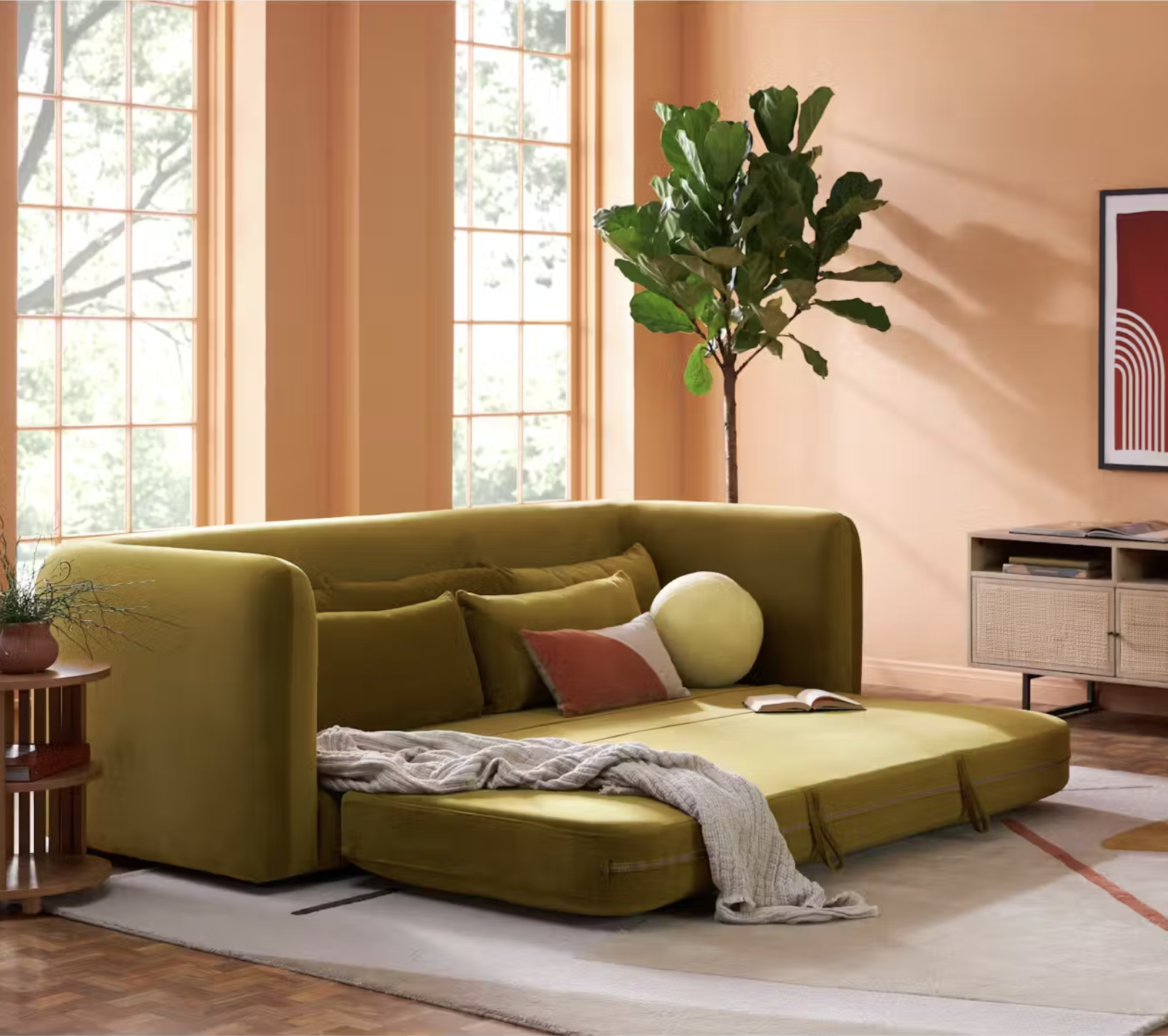 A green sofa bed with cushions and a blanket is set up in a sunlit living room next to a large plant, wooden table, and cabinet with framed wall art.