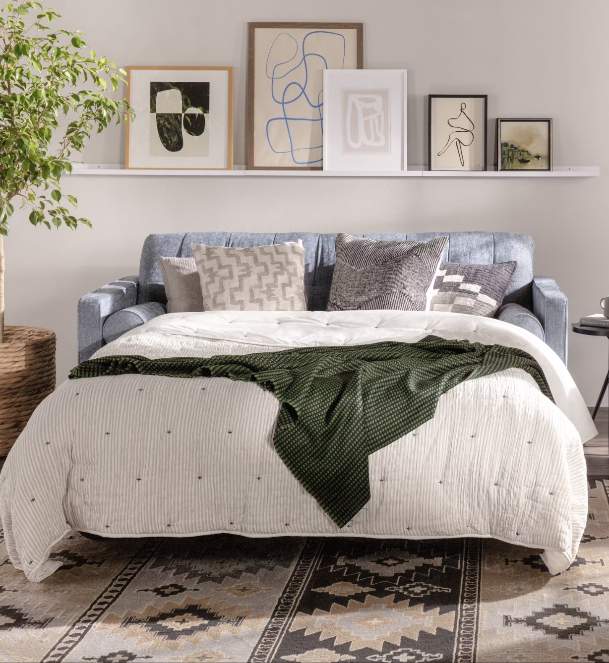 A sofa bed with a light quilt and dark green throw sits in a cozy room, accented by geometric pillows, wall art, a potted plant, and a patterned rug.