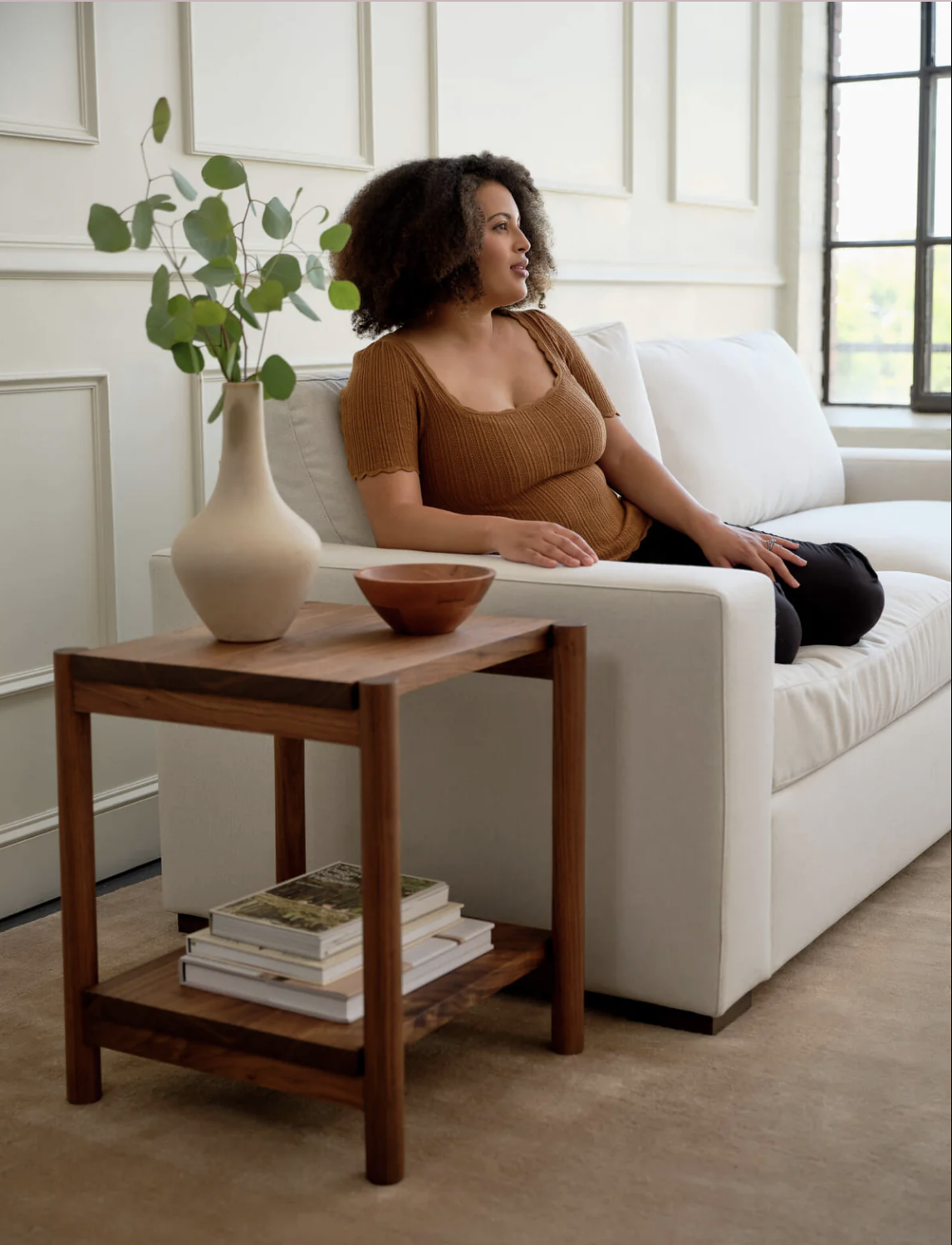 A woman sits on a white sofa next to a wooden side table with a vase, bowl, and books in a bright, modern living room.