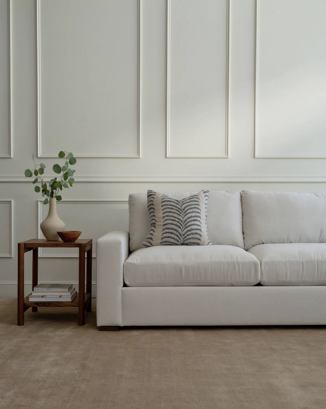 A white sofa with a patterned cushion sits next to a wooden side table holding a vase with greenery and a small bowl, against a paneled white wall.