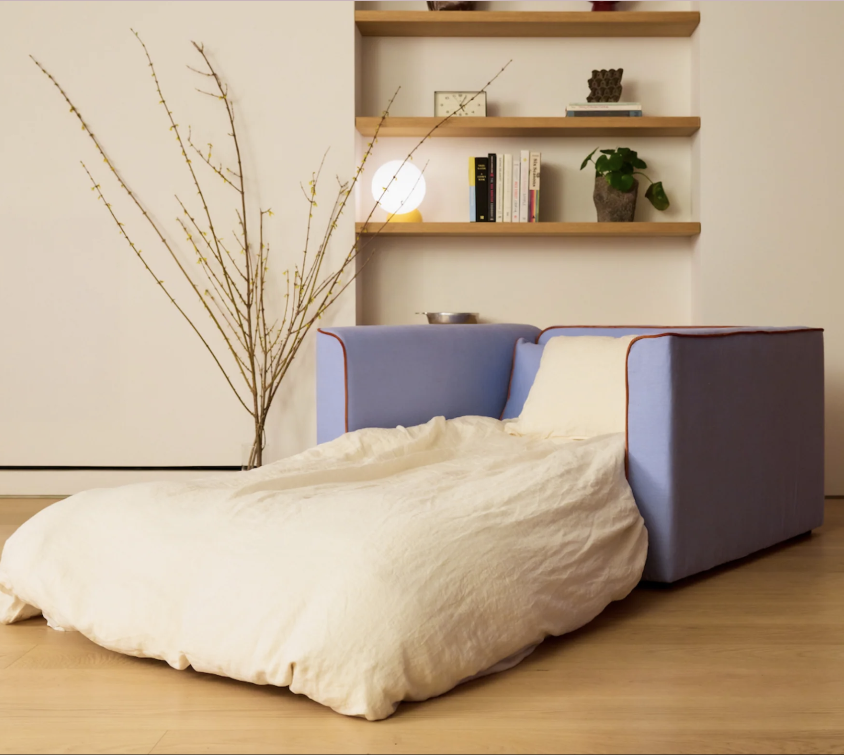 A blue armchair with a cream-colored blanket or cover extends into a low, padded lounge seat; shelves and a bare branch decor are in the background.