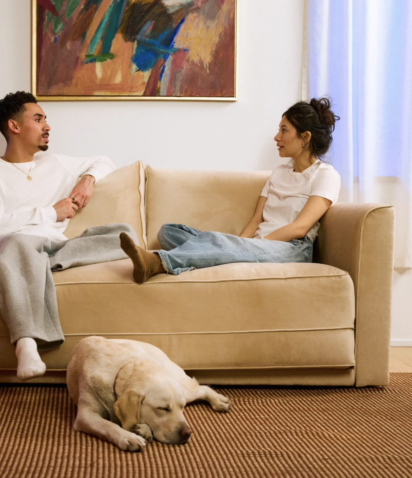 Two people sit on a beige couch having a conversation, while a yellow labrador dog lies on the carpeted floor in front of them.