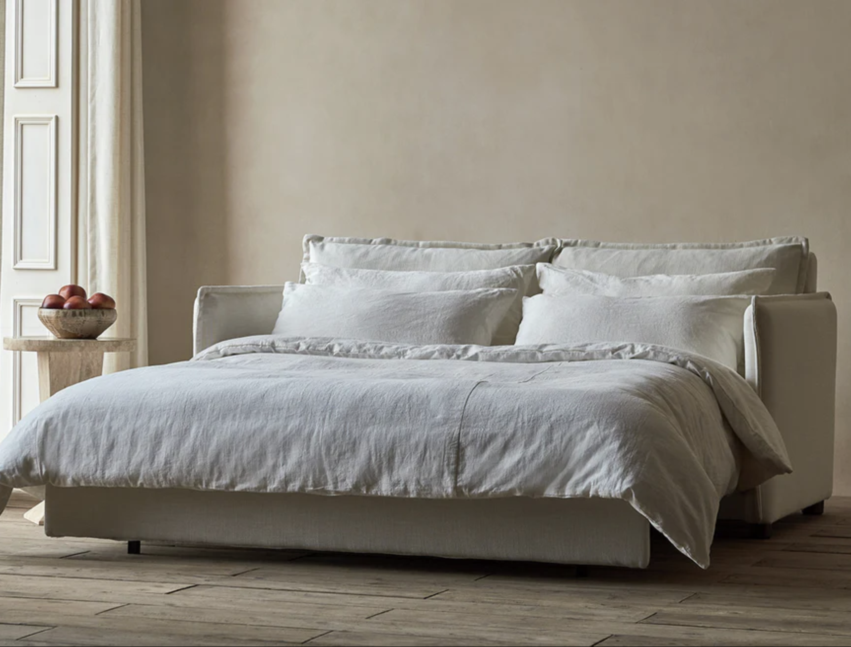 A cream-colored sofa bed is extended with white bedding and pillows, placed beside a round wooden side table with a bowl of apples in a minimally decorated room.