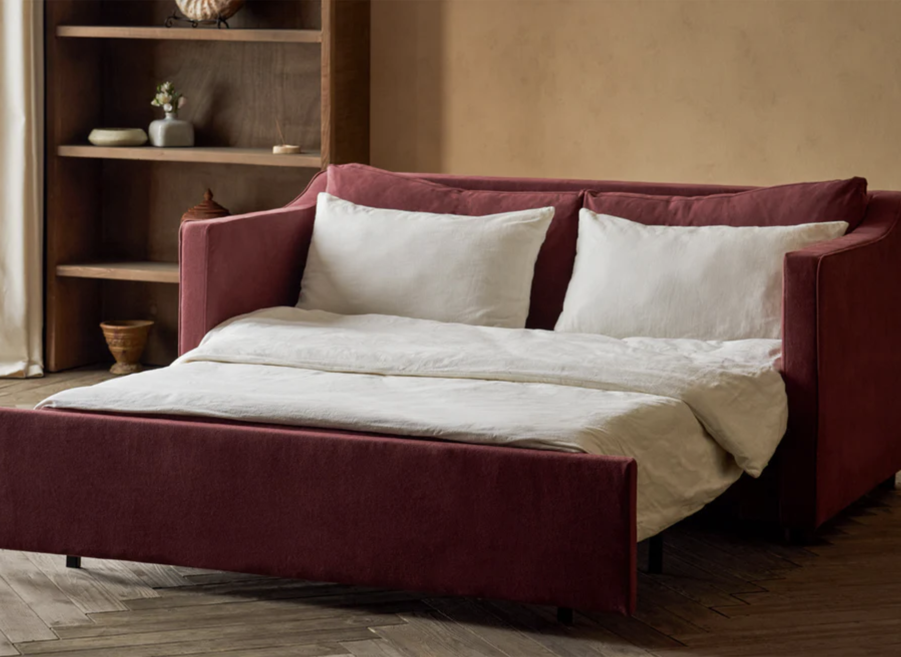 A burgundy upholstered sleeper sofa is unfolded into a bed with white bedding and pillows in a room with wooden shelves and a beige wall.