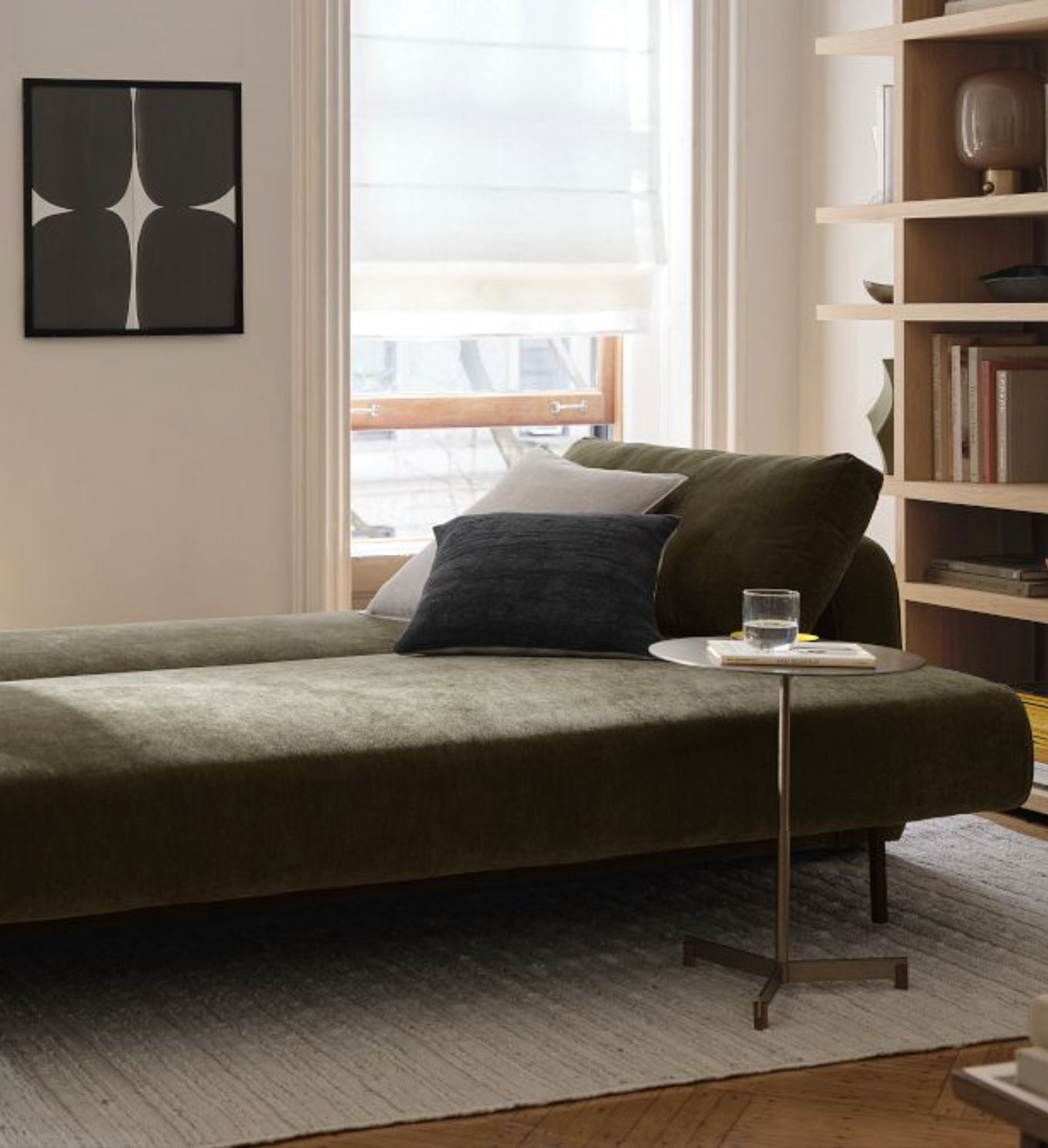 A green velvet daybed with two pillows sits beside a round side table with a glass and book, in a bright room with shelves, art, and large windows.