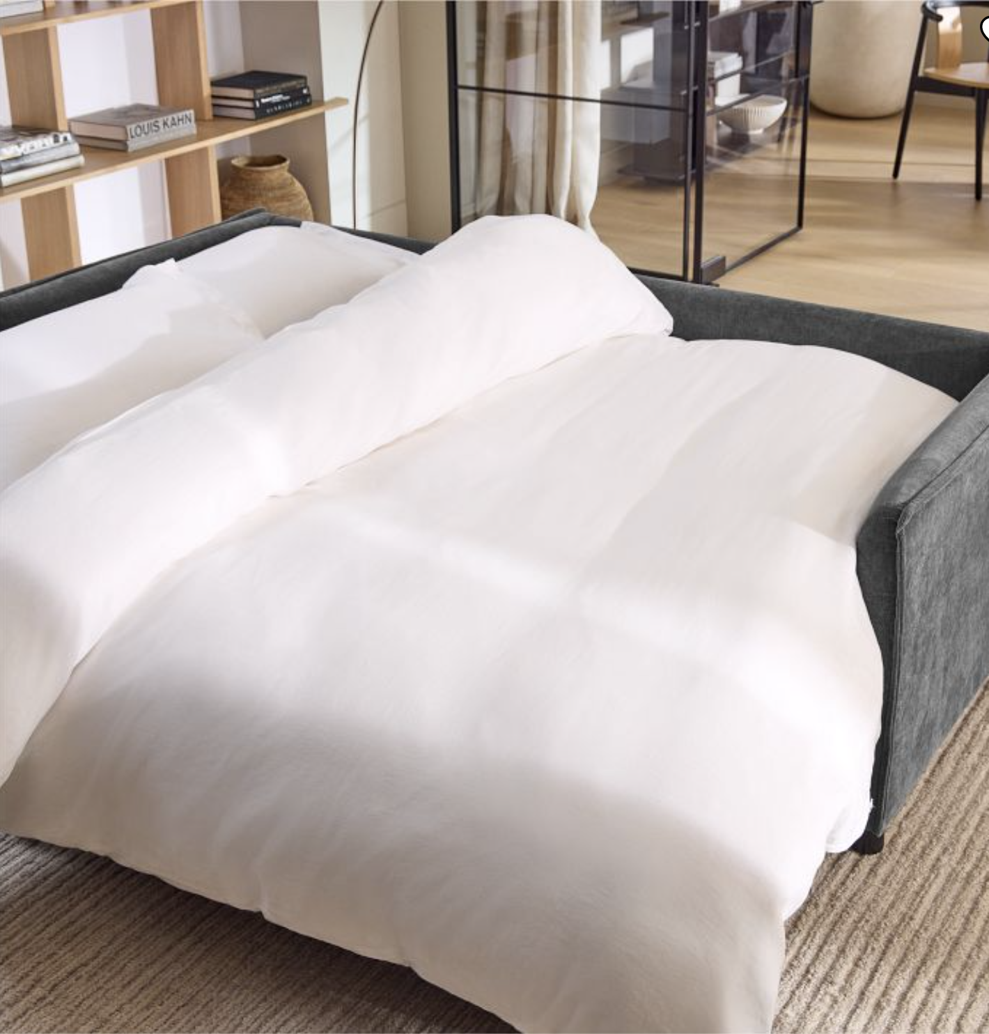 A gray sofa bed is unfolded with white bedding on it, placed on a beige rug in a modern living room with shelves and glass doors in the background.
