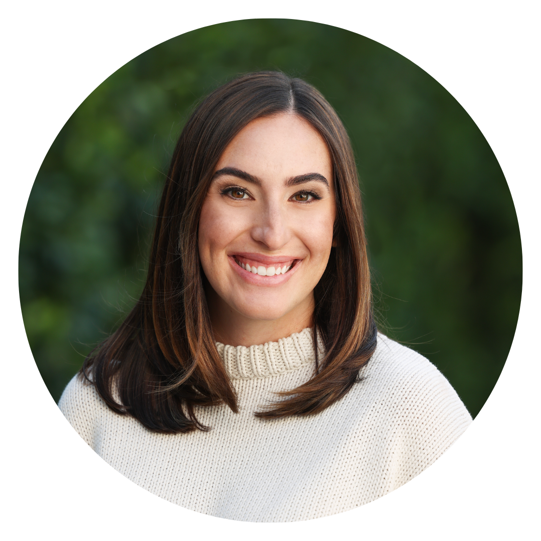 A woman with straight brown hair and a white sweater smiles at the camera, with a blurred green background.